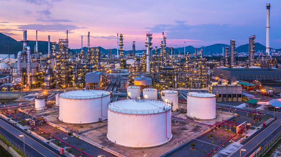 This image captures a stunning aerial view of a large petrochemical refinery complex during dusk, featuring white storage tanks in the foreground and towering industrial structures in the background illuminated by a colorful sunset.