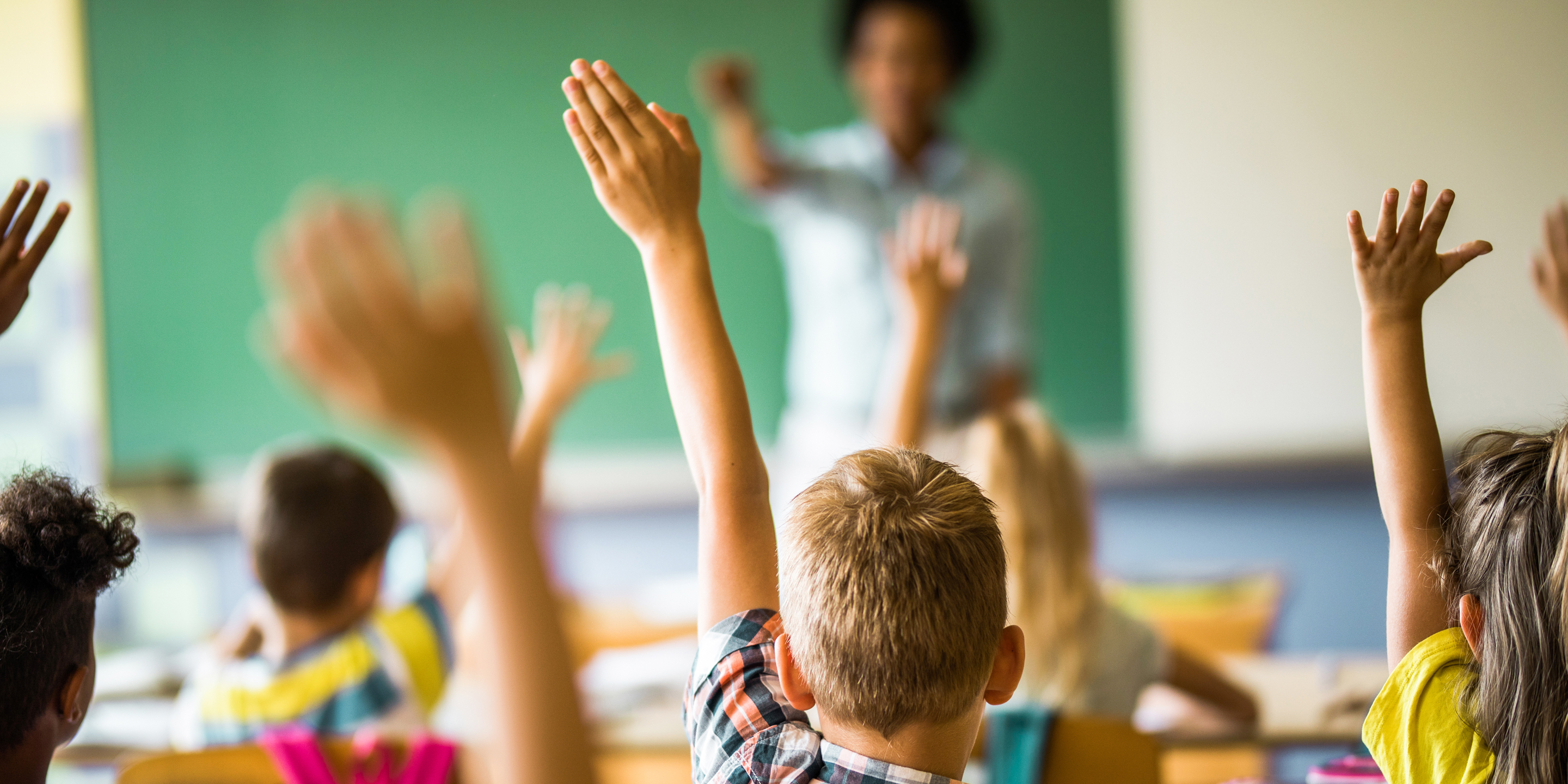 Rear view of large group of students raising their hands to answer the question on a class at elementary school.