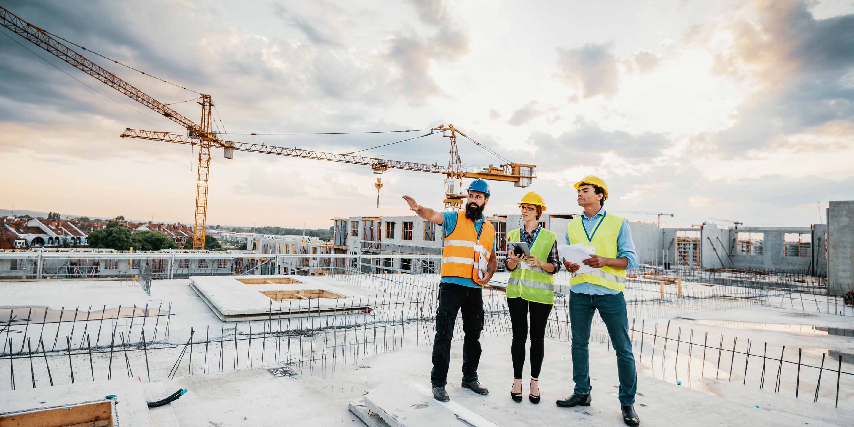 Three construction workers in safety vests and helmets stand on a construction site, with one pointing into the distance and others holding documents. Cranes and partially built structures are visible in the background under a partly cloudy sky.