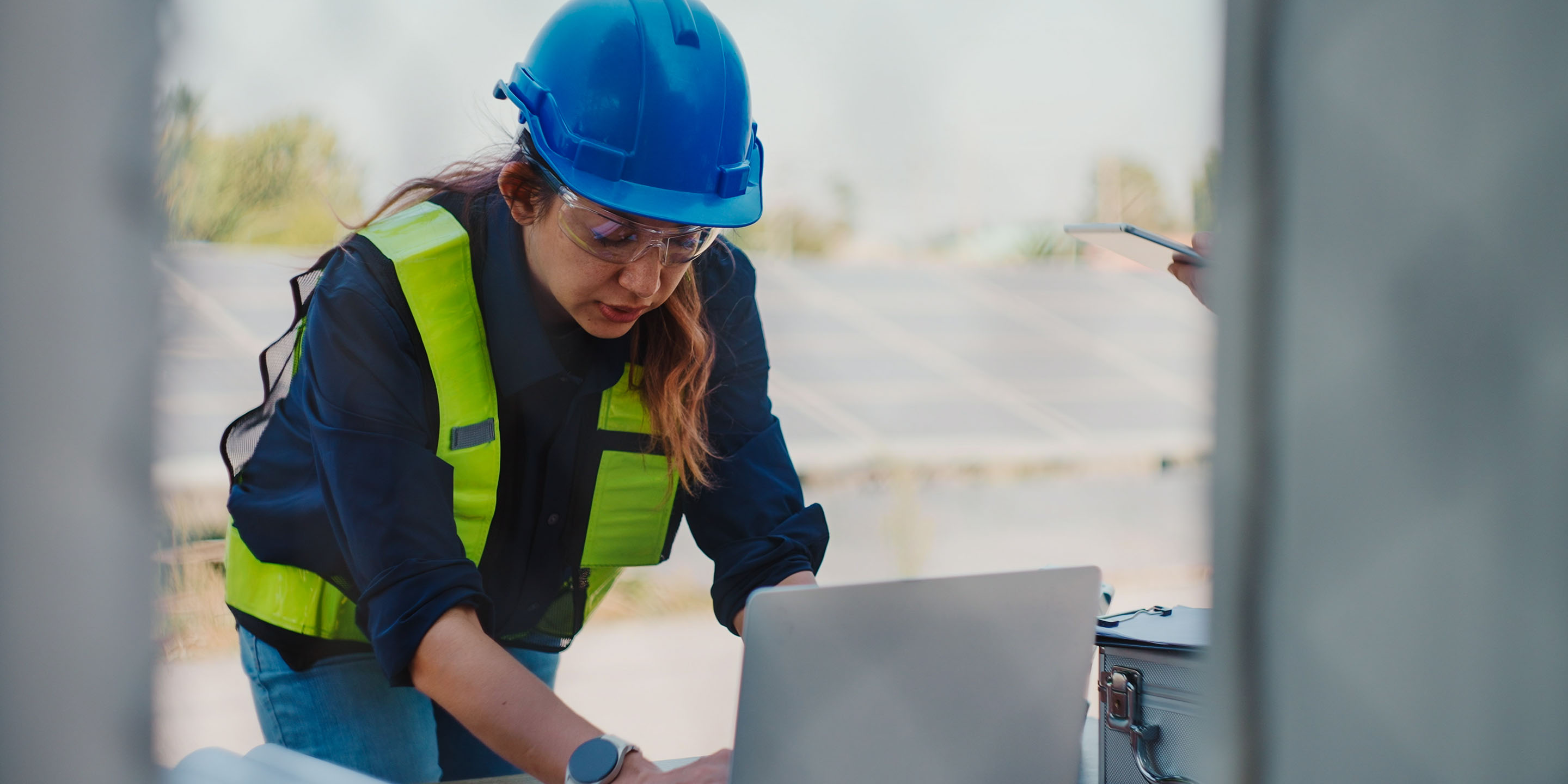 A person in a blue hard hat and high-visibility vest works on a laptop at an outdoor industrial site with solar panels. Another individual is partially visible, holding a tablet or clipboard, highlighting technology use in sustainable construction.