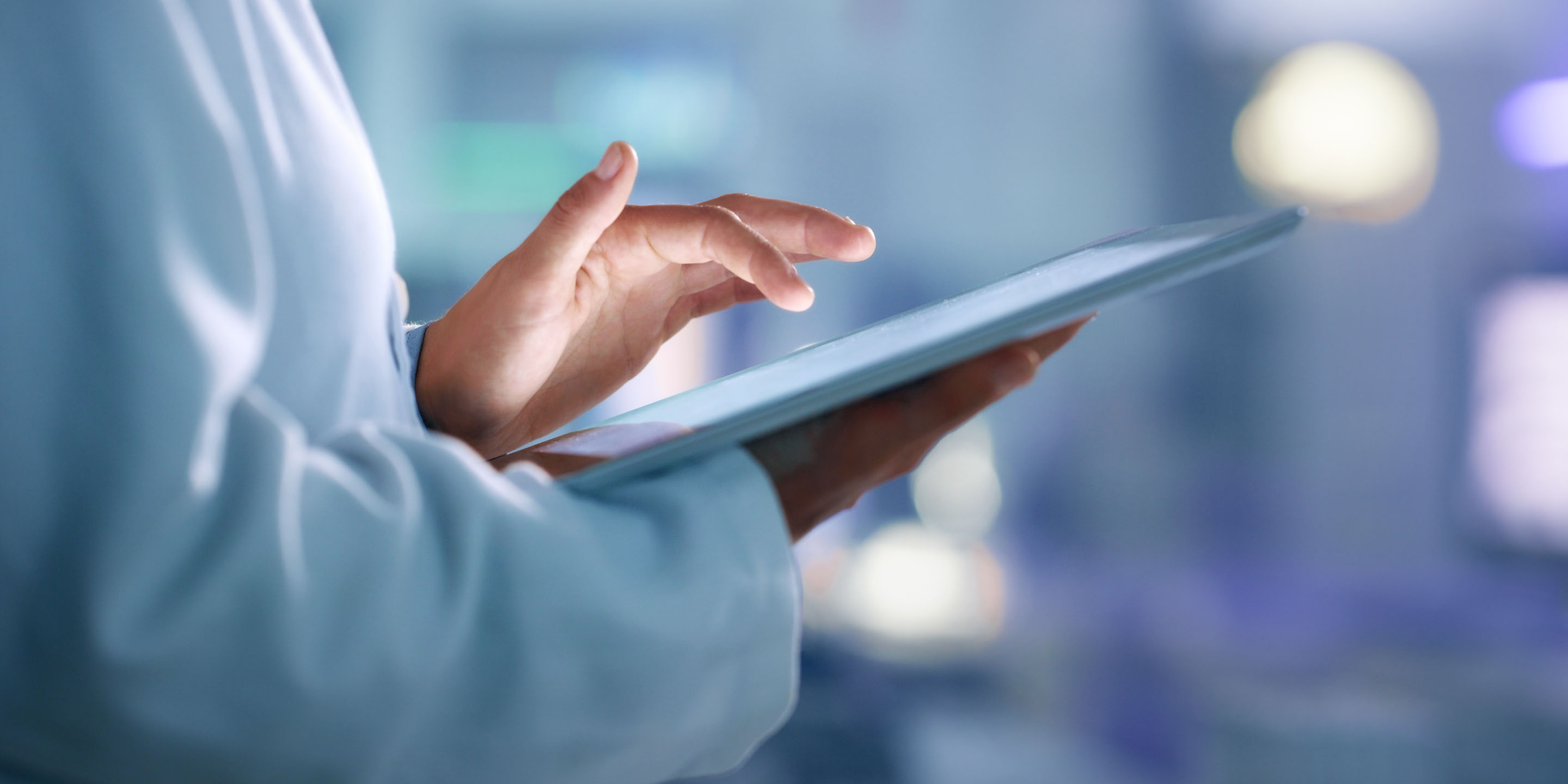 A person in a white lab coat interacts with a tablet device in a blurred indoor environment, suggesting a medical or laboratory setting. The image highlights the integration of technology in healthcare or research.