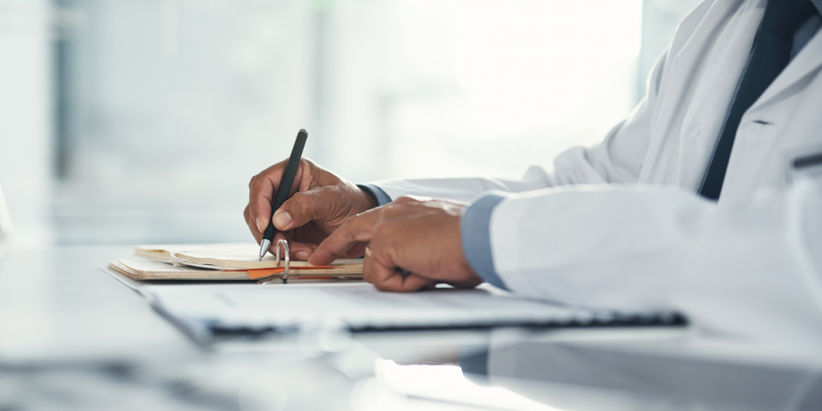 A close-up of a medical professional in a white coat writing on a clipboard or notepad. The desk includes documents that appear to be medical records, emphasizing clinical documentation and healthcare practice.