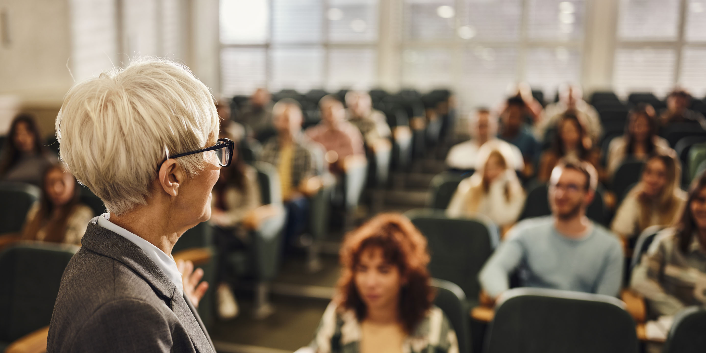 A classroom or lecture hall scene featuring a professor or lecturer standing at the front, speaking to an audience of students. The image captures the academic environment with natural light filtering through windows and students attentively facing forward.