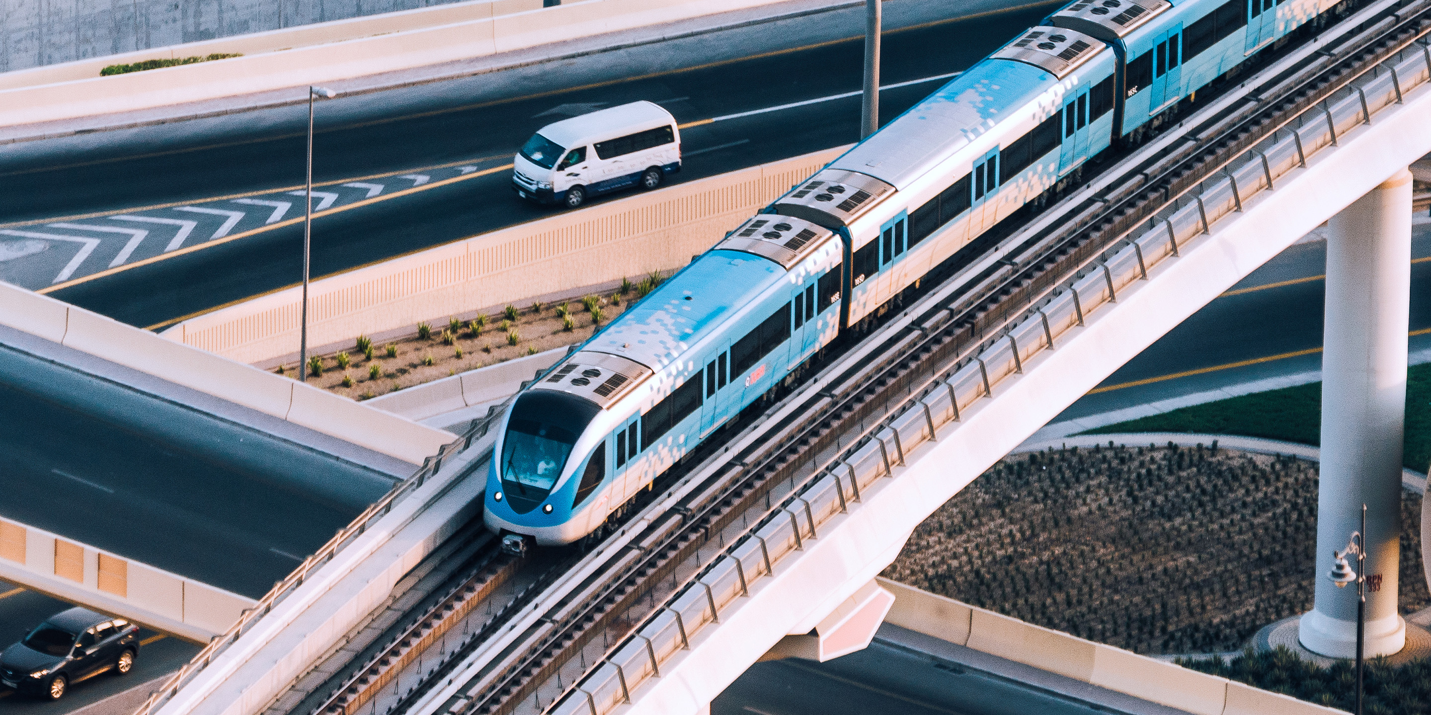 A sleek blue and white elevated train crosses a concrete bridge above a busy road with a white van below. Ideal for showcasing urban transportation, infrastructure, or smart city mobility solutions.