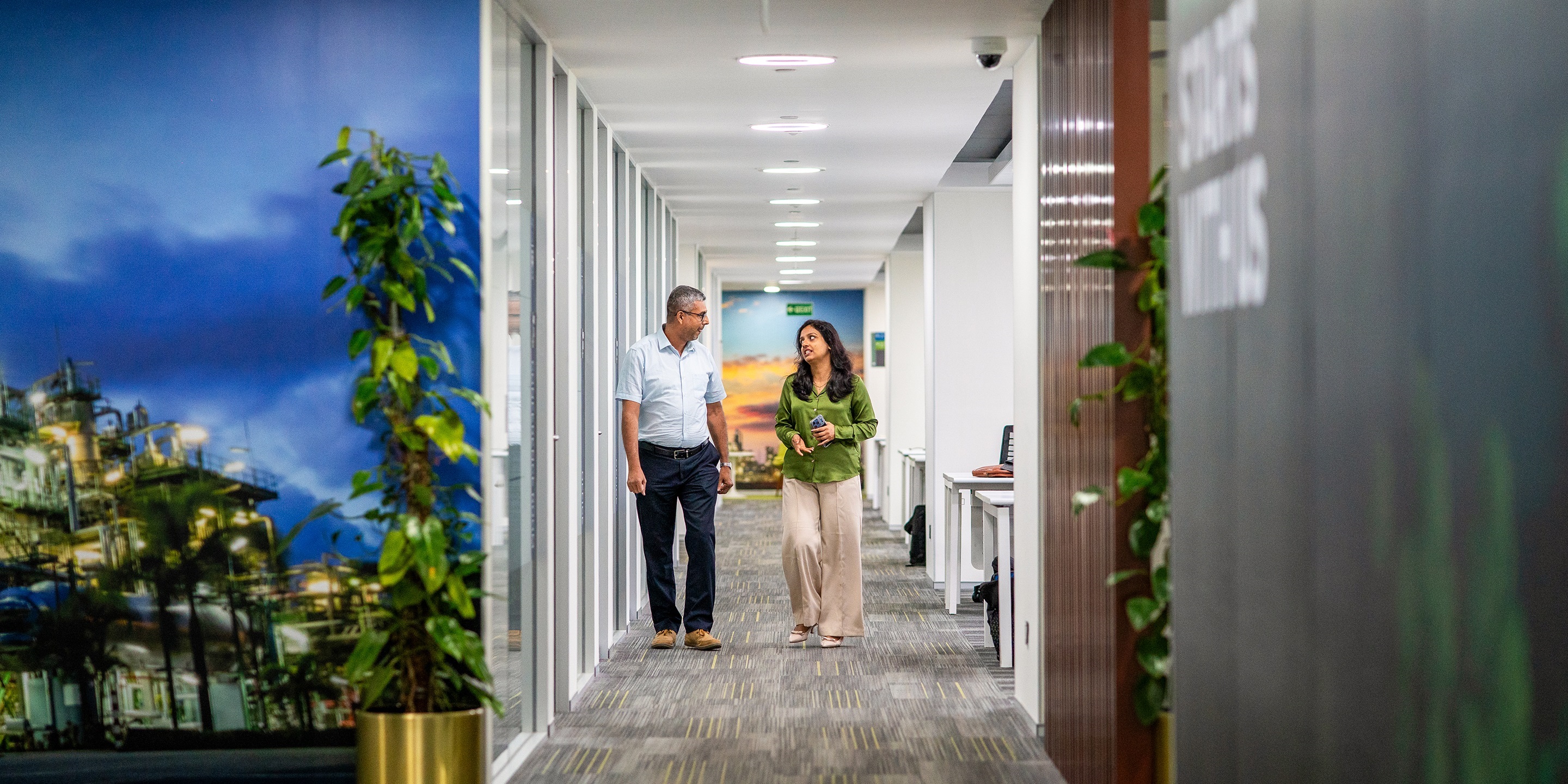 Professionals walking and talking in a modern office corridor with clean design and greenery.