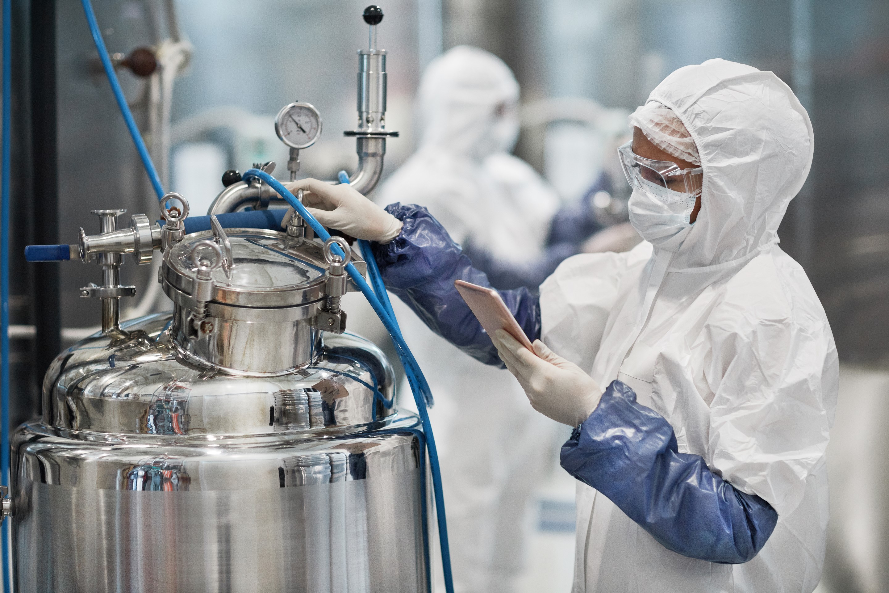 A lab technician in protective clothing checks equipment while holding a tablet in a high-tech facility.