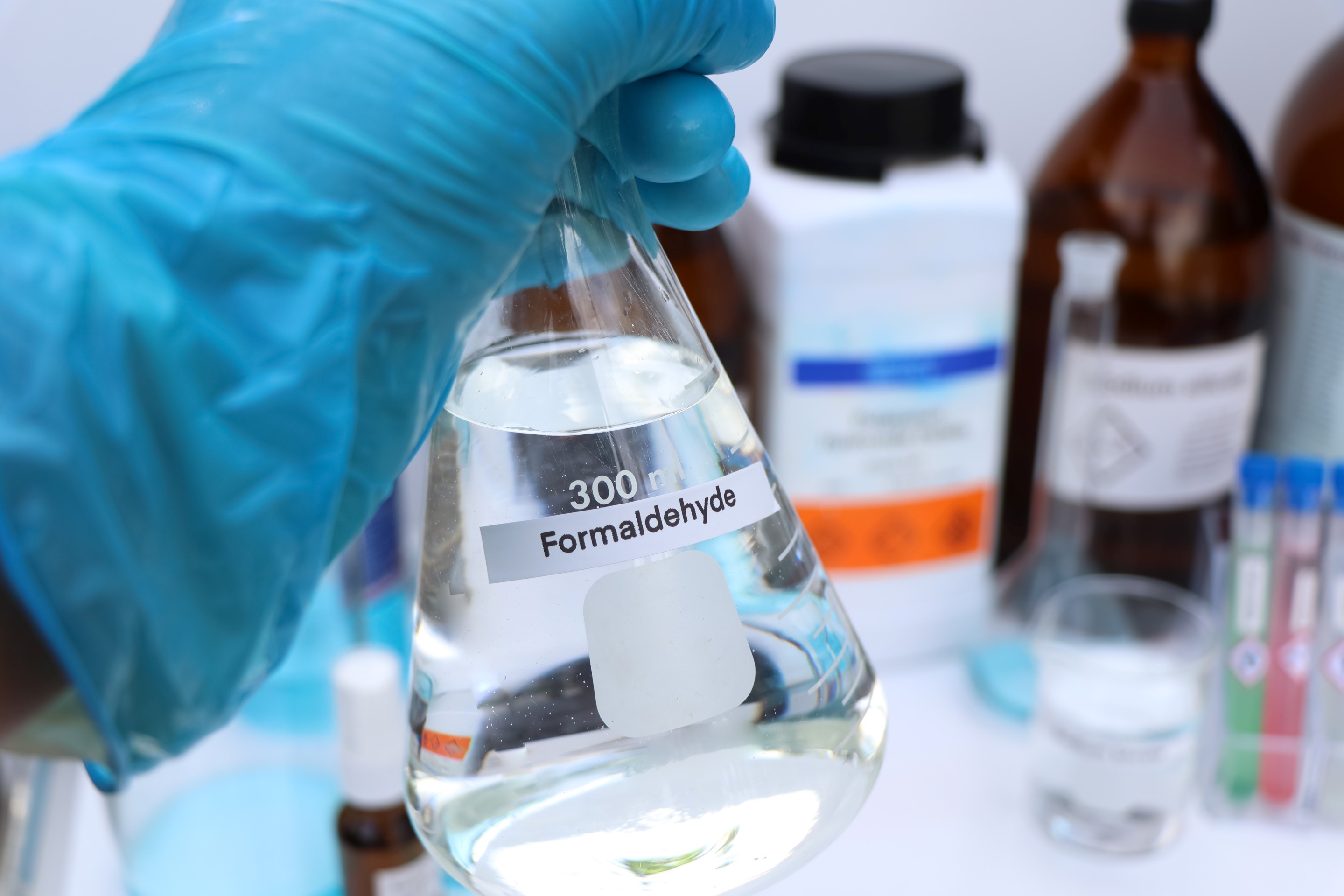 A lab technician in blue gloves holds a labeled flask of formaldehyde, surrounded by various lab materials.