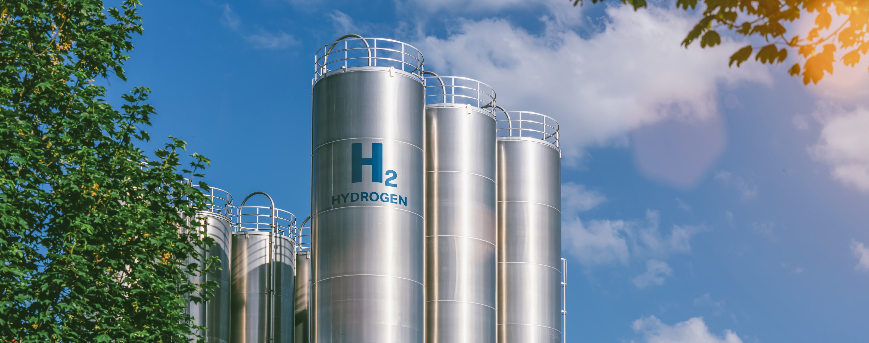 A view of tall hydrogen storage tanks marked with "H2," framed by a bright sky and greenery.