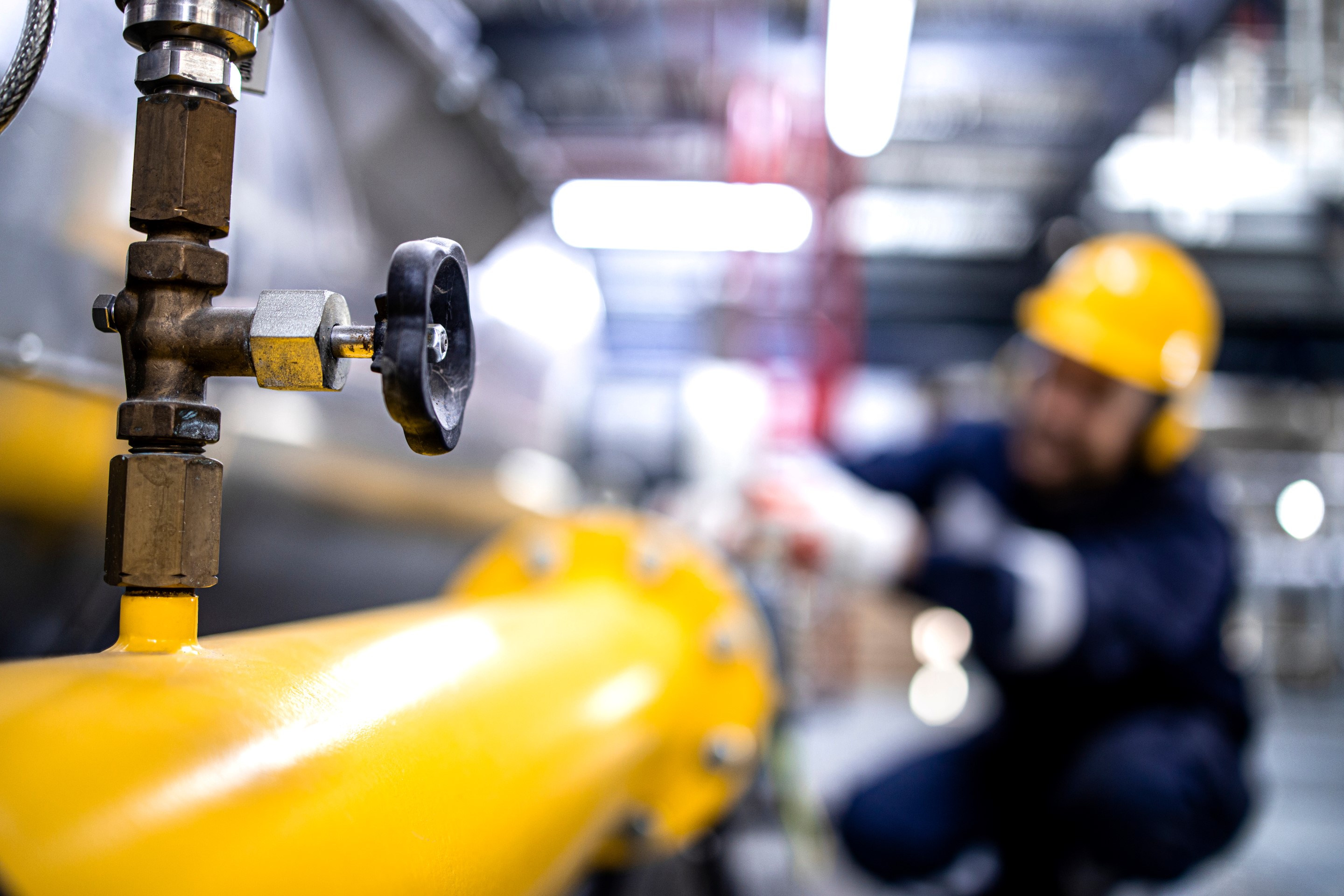 Detailed shot of a valve on a yellow pipe, with an industrial worker visible in the background.