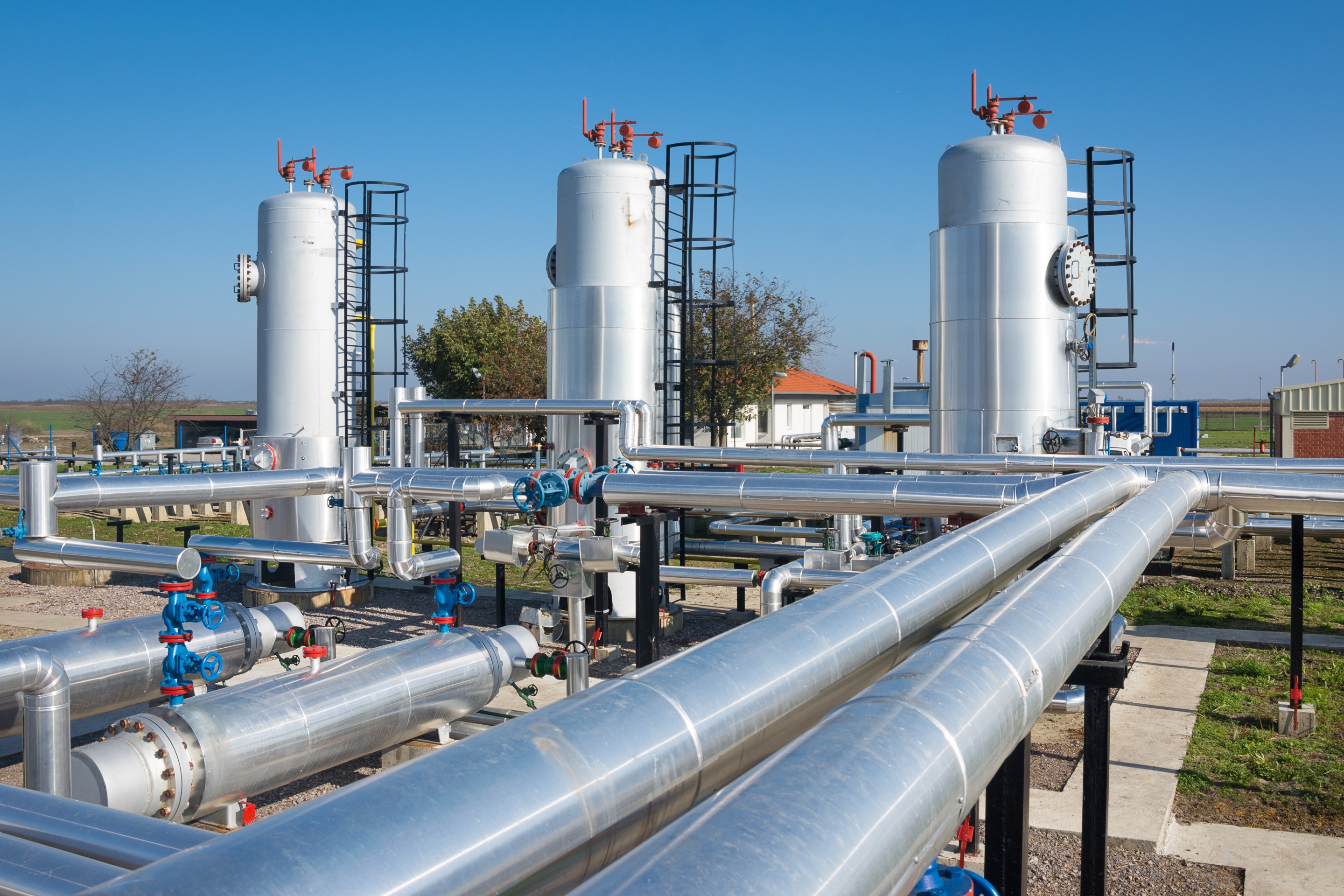 A gas processing facility featuring shiny pipelines, large storage tanks, and a clear blue sky in the background.