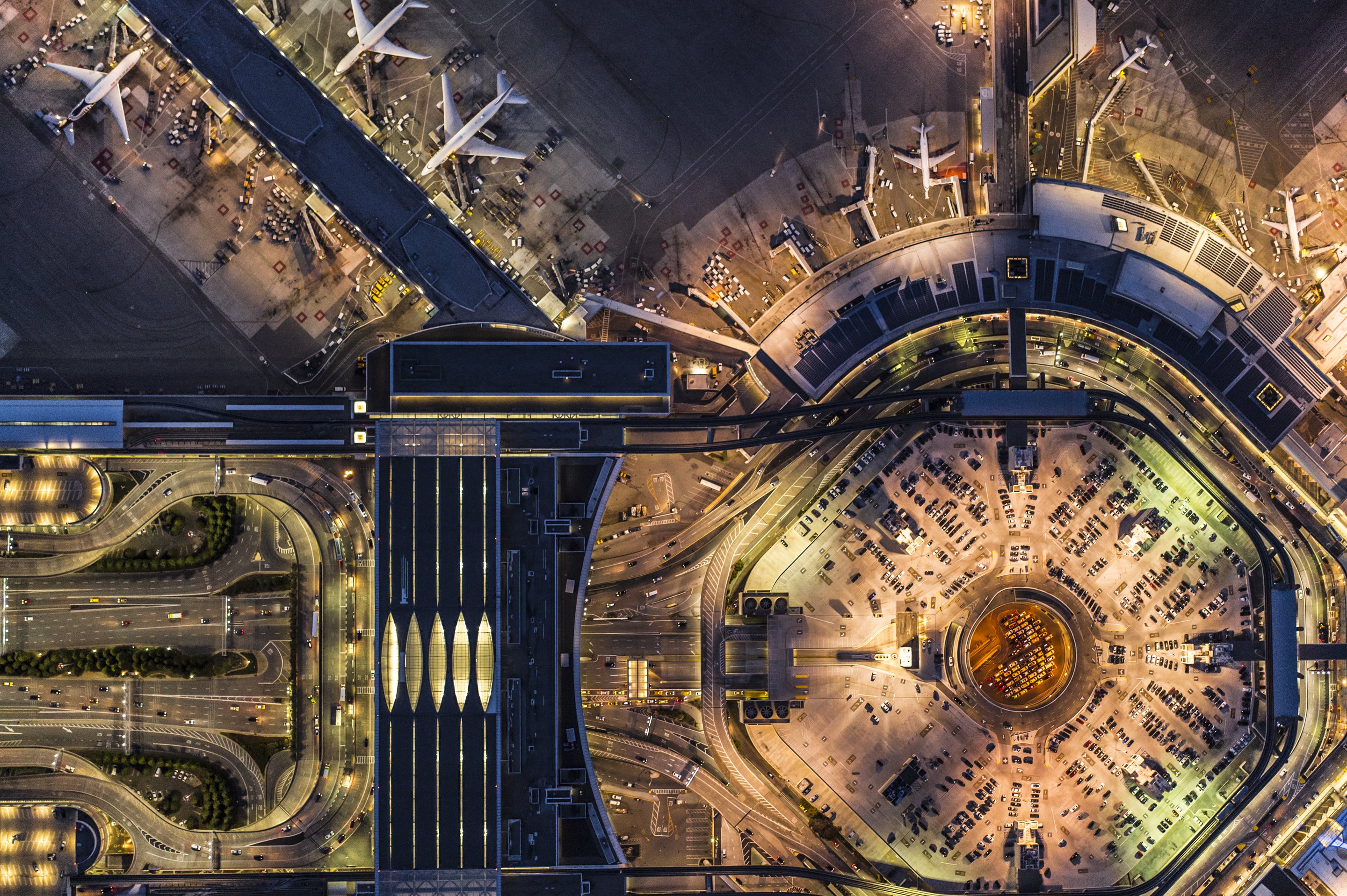An illuminated aerial view capturing the intricate night-time layout of airport terminals, aircraft parking aprons, and multi-level traffic infrastructure.