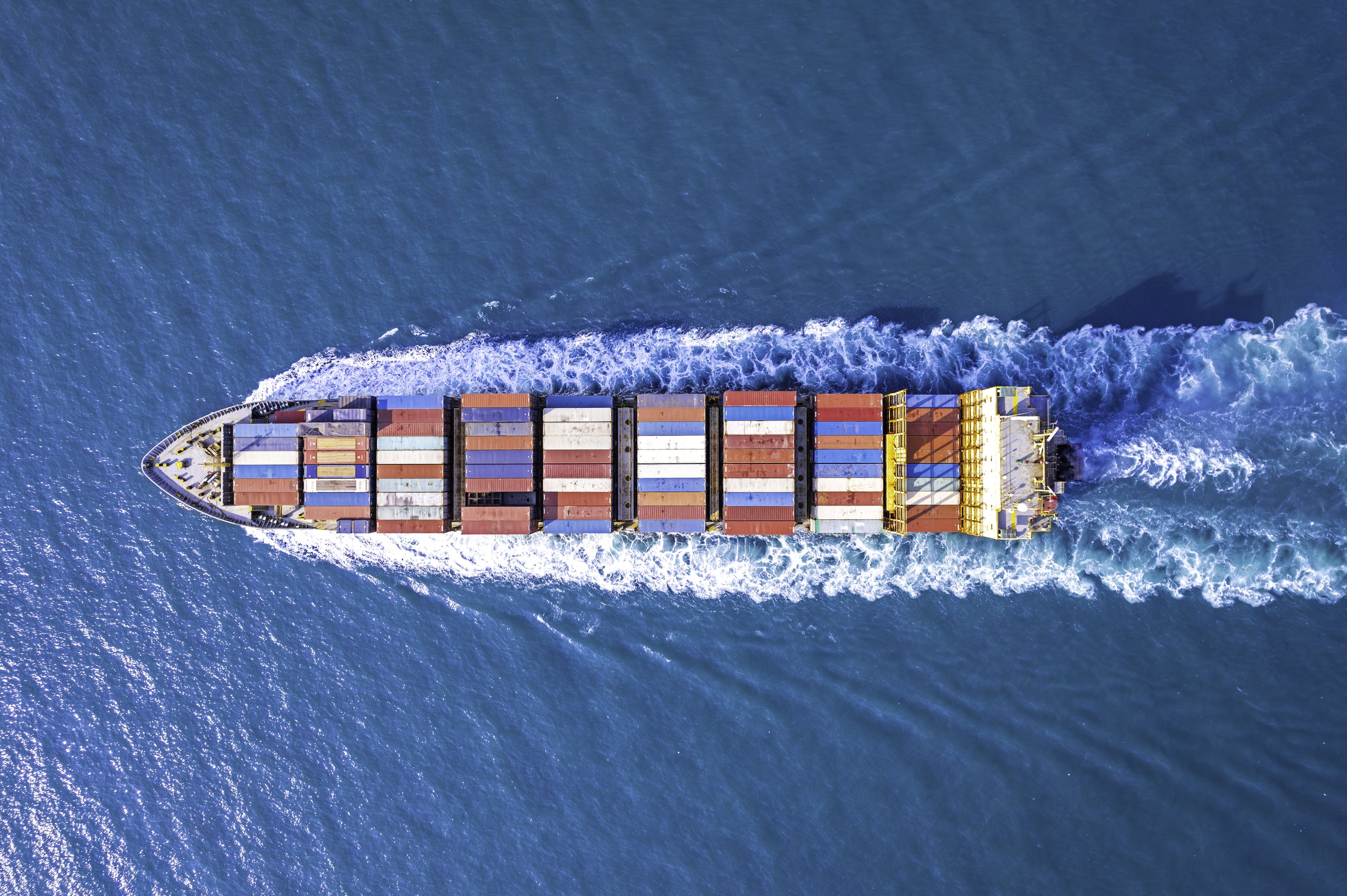 Aerial view of a massive container ship traversing the open ocean while creating a white wake in deep blue water.