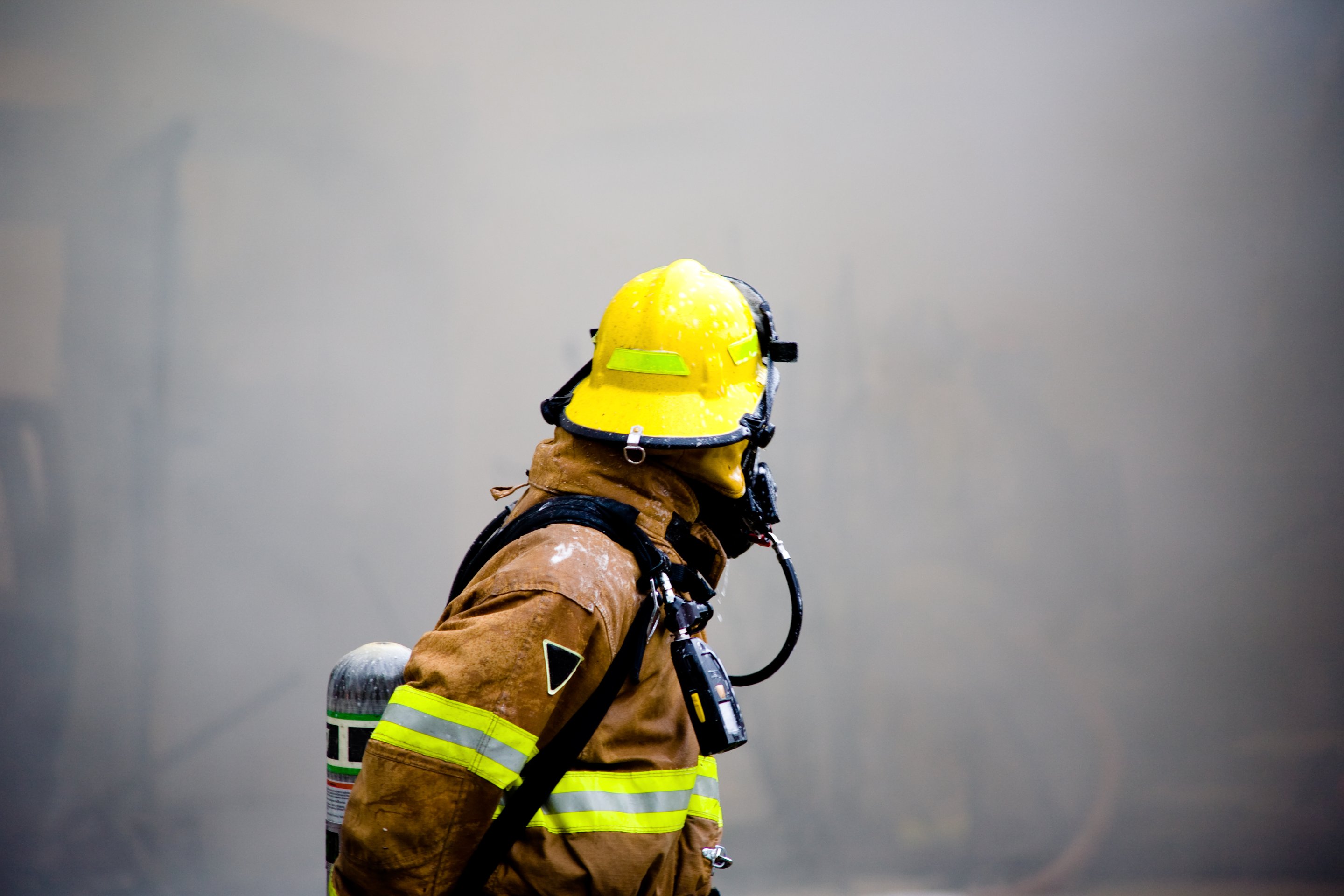 A firefighter in full protective gear and helmet navigates through dense, hazy smoke at the scene of an emergency.