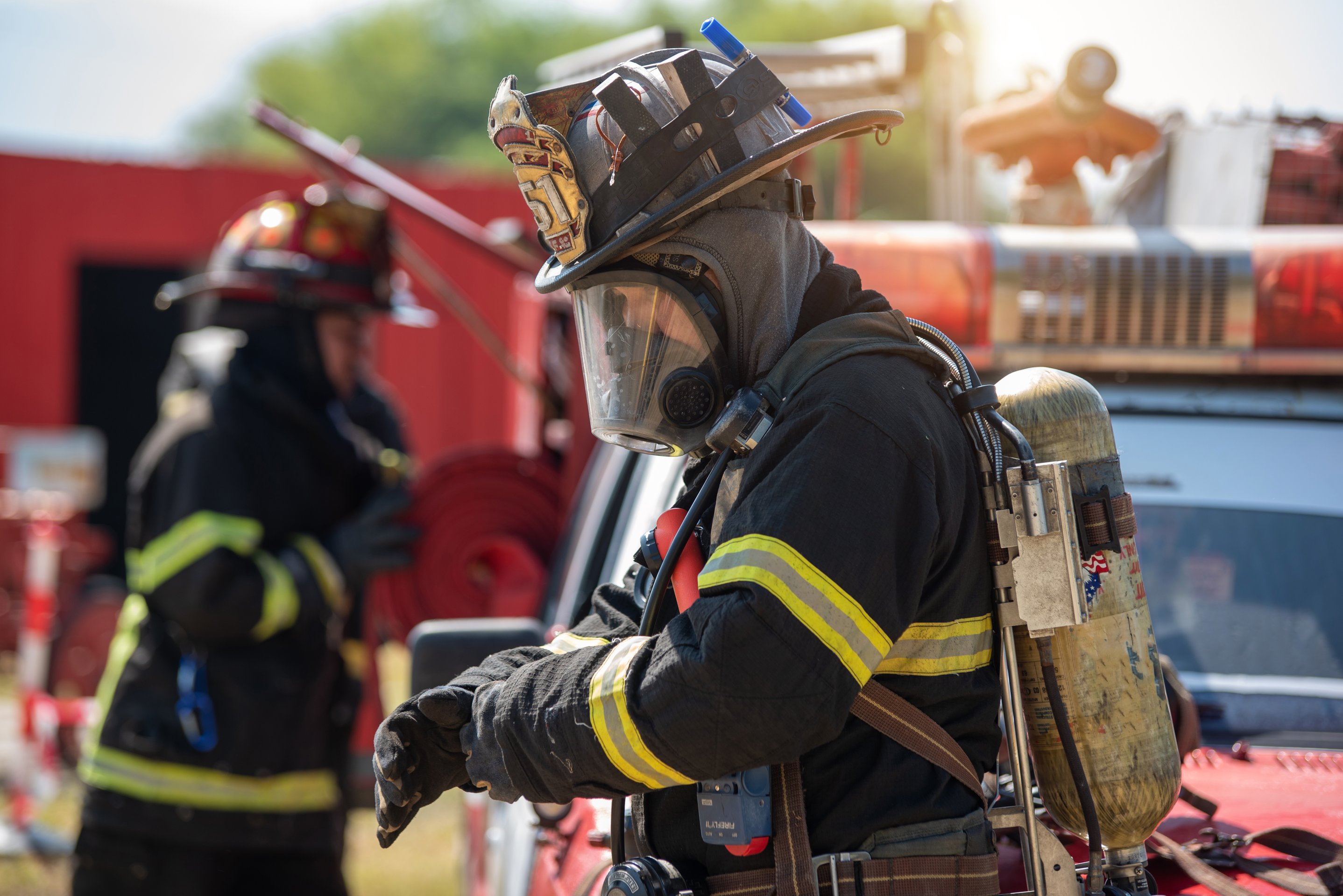 A professional firefighter in full protective turnout gear and SCBA respirator prepares for duty near a fire engine on an active training site.