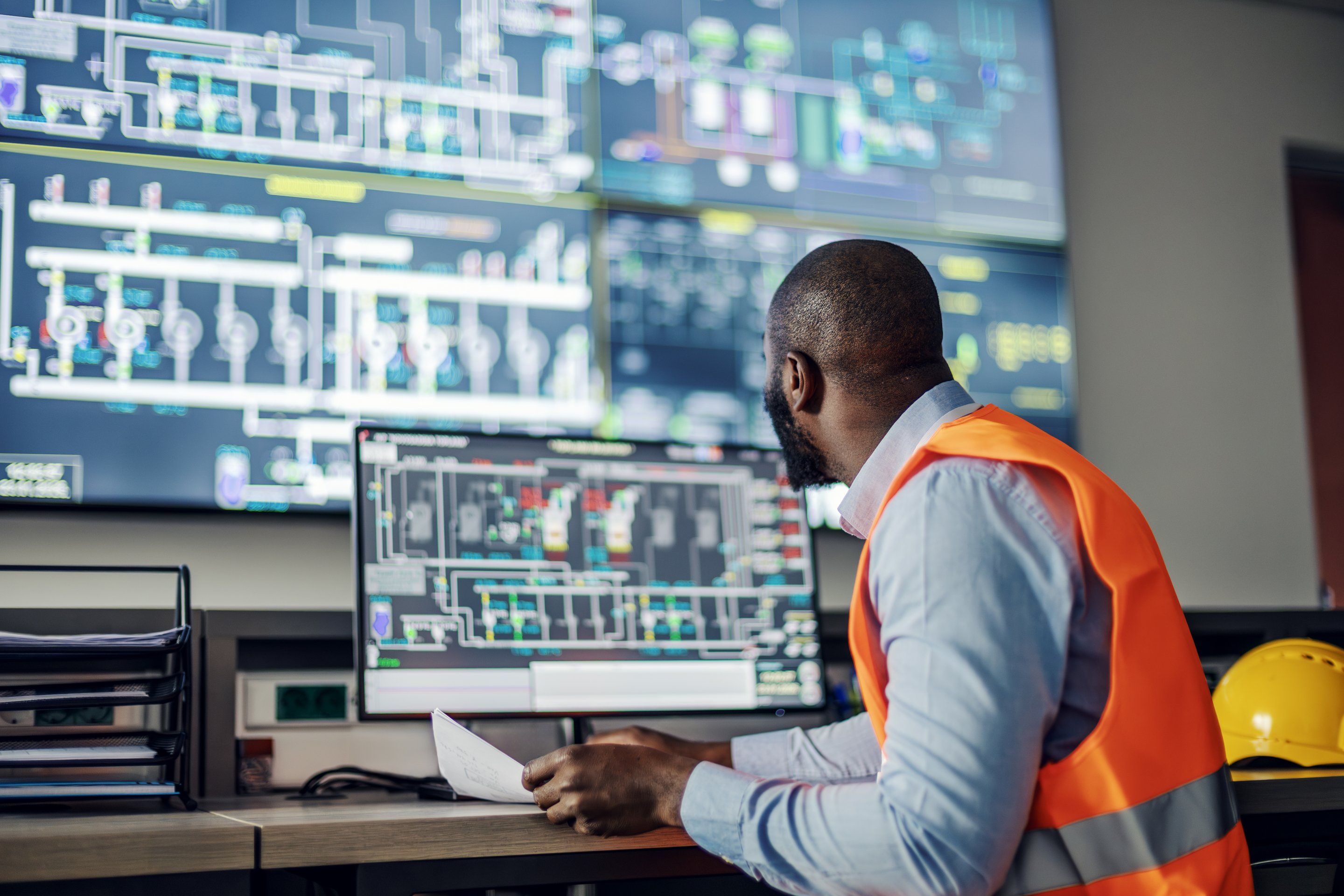 An engineer monitors complex infrastructure data on multiple digital screens while working in a high-tech facility control center.