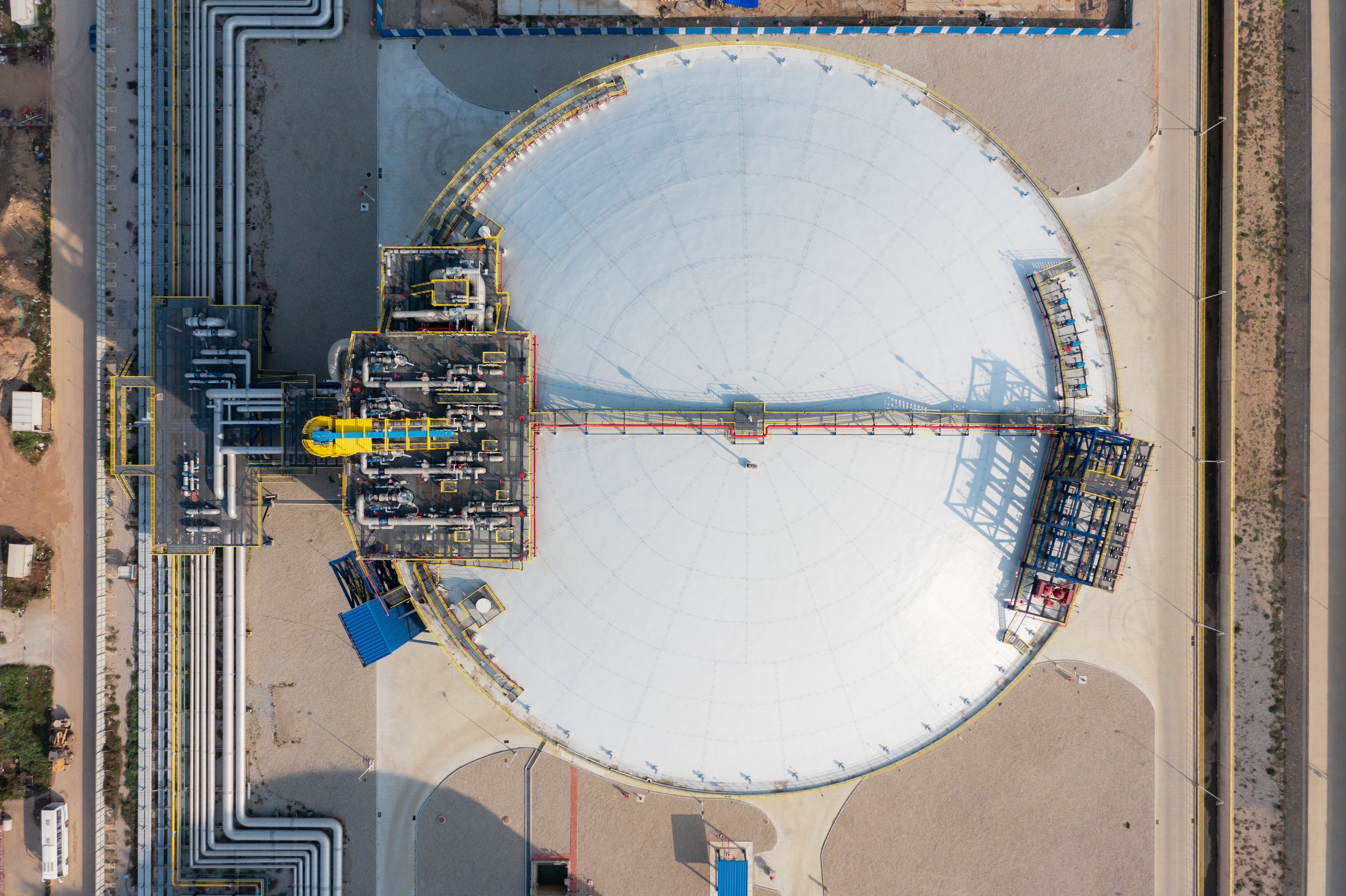 Aerial view of a large white cylindrical fuel storage tank connected to complex industrial piping infrastructure.