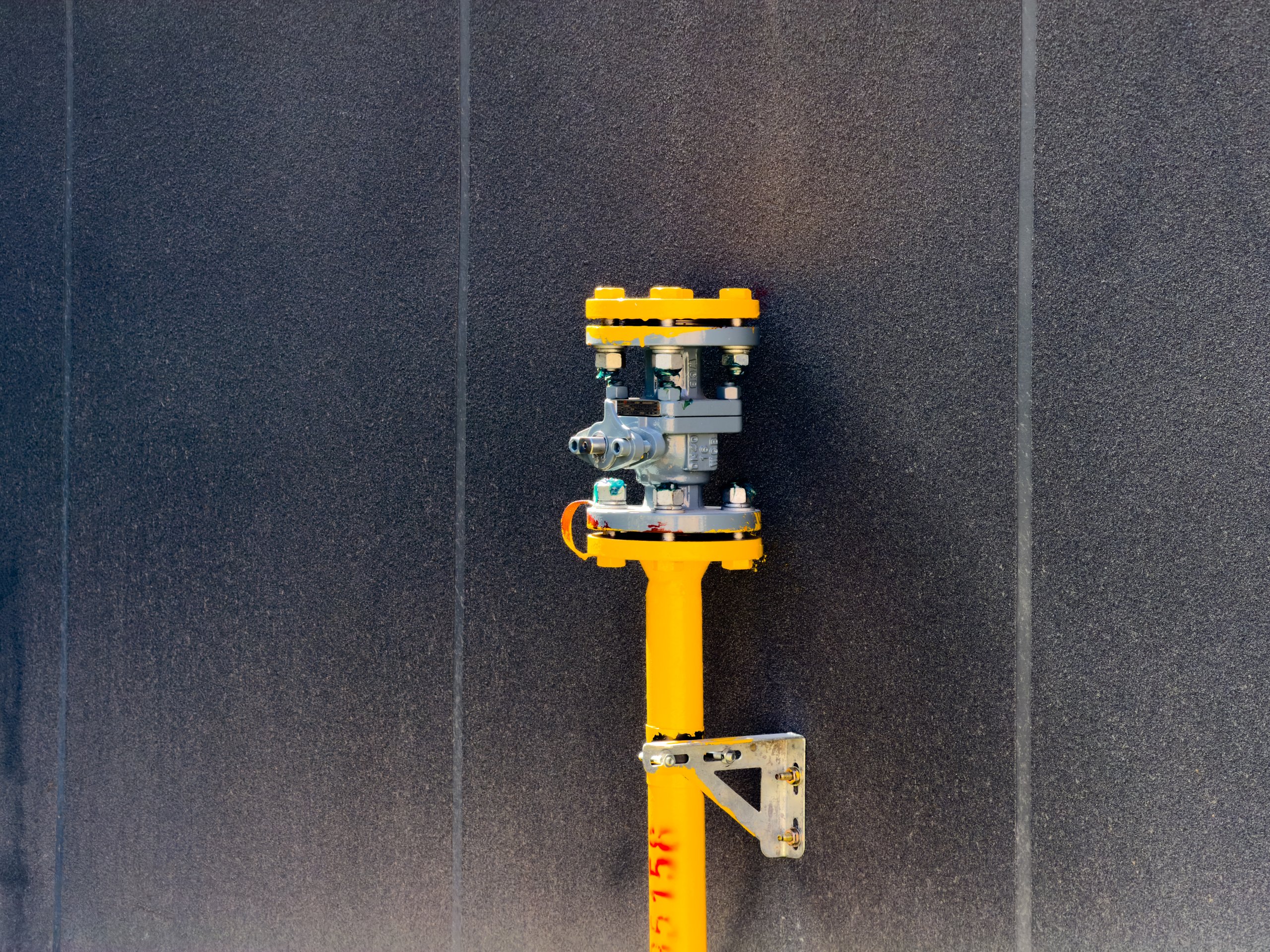 A bright yellow industrial valve stands mounted against a modern, textured dark grey wall.