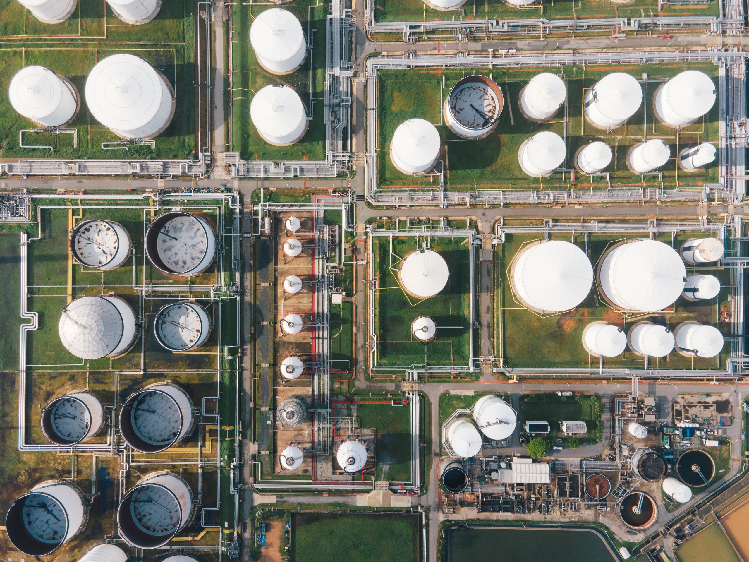 An aerial view displays an industrial refinery with numerous storage tanks and a complex network of interconnected pipelines.