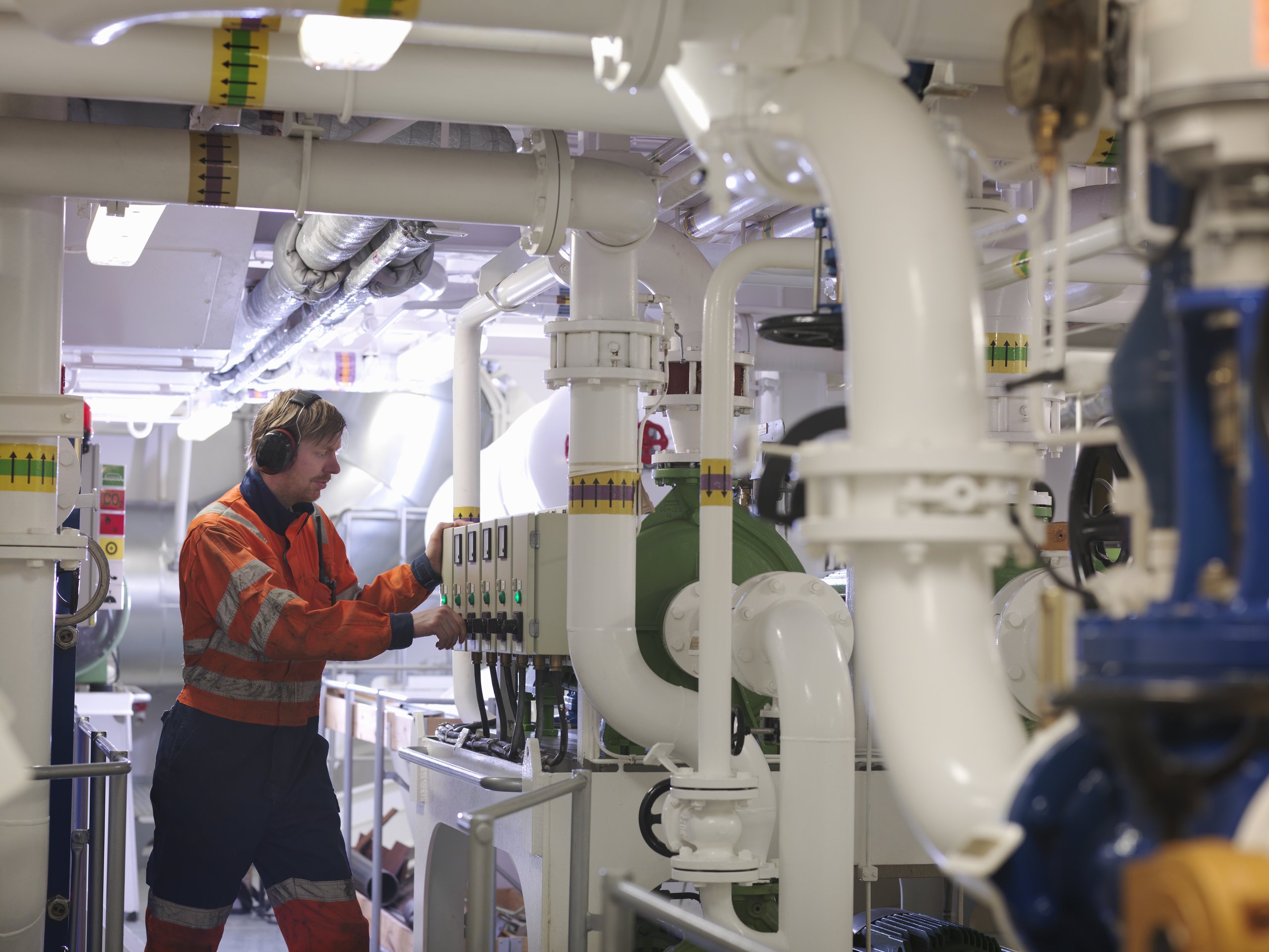 An industrial technician in high-visibility workwear operates machinery controls amidst a complex network of white pipes in a maritime engine room.