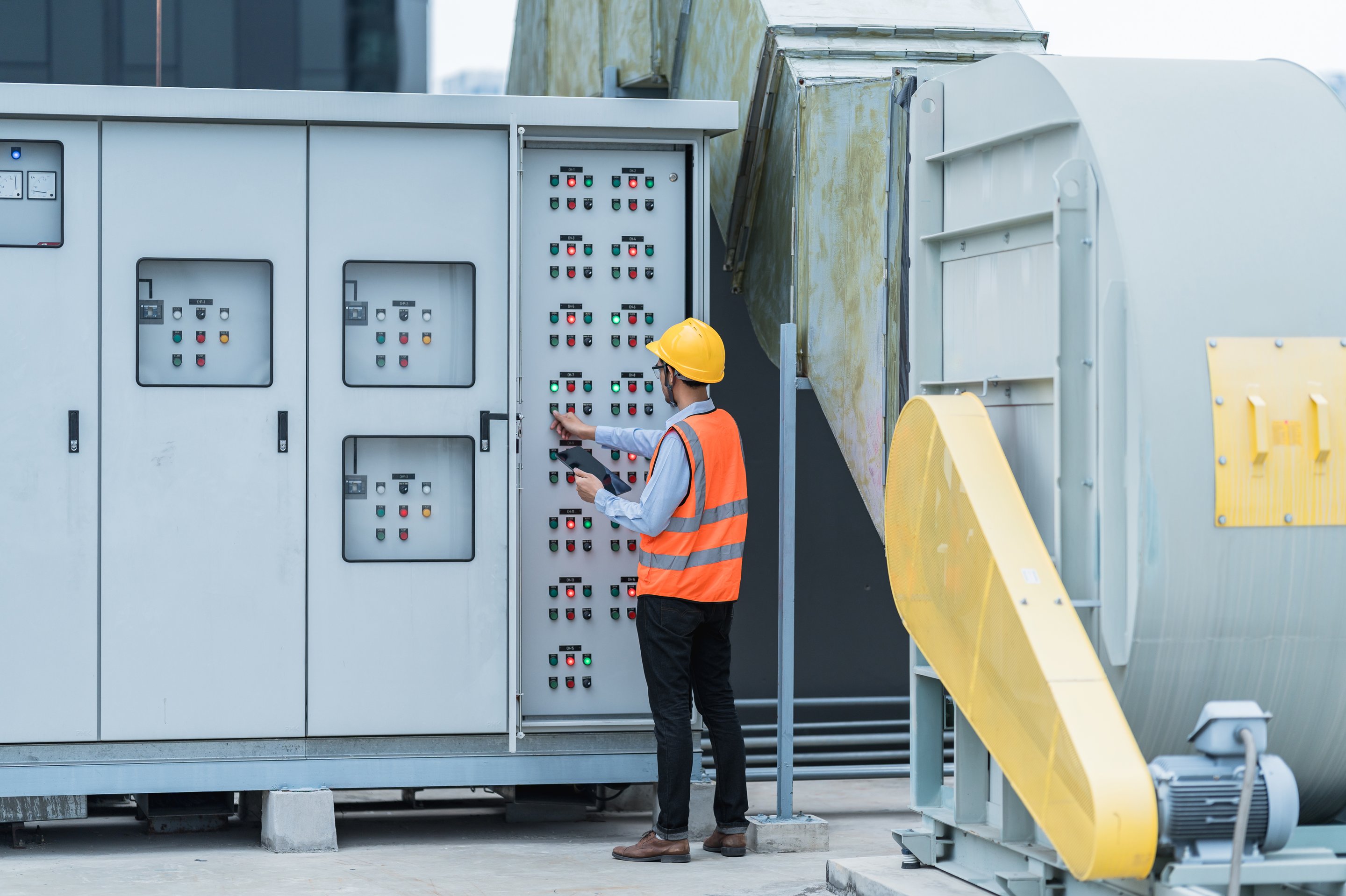 A technician in a hard hat and high-visibility vest inspects and operates an industrial control panel on a rooftop unit.