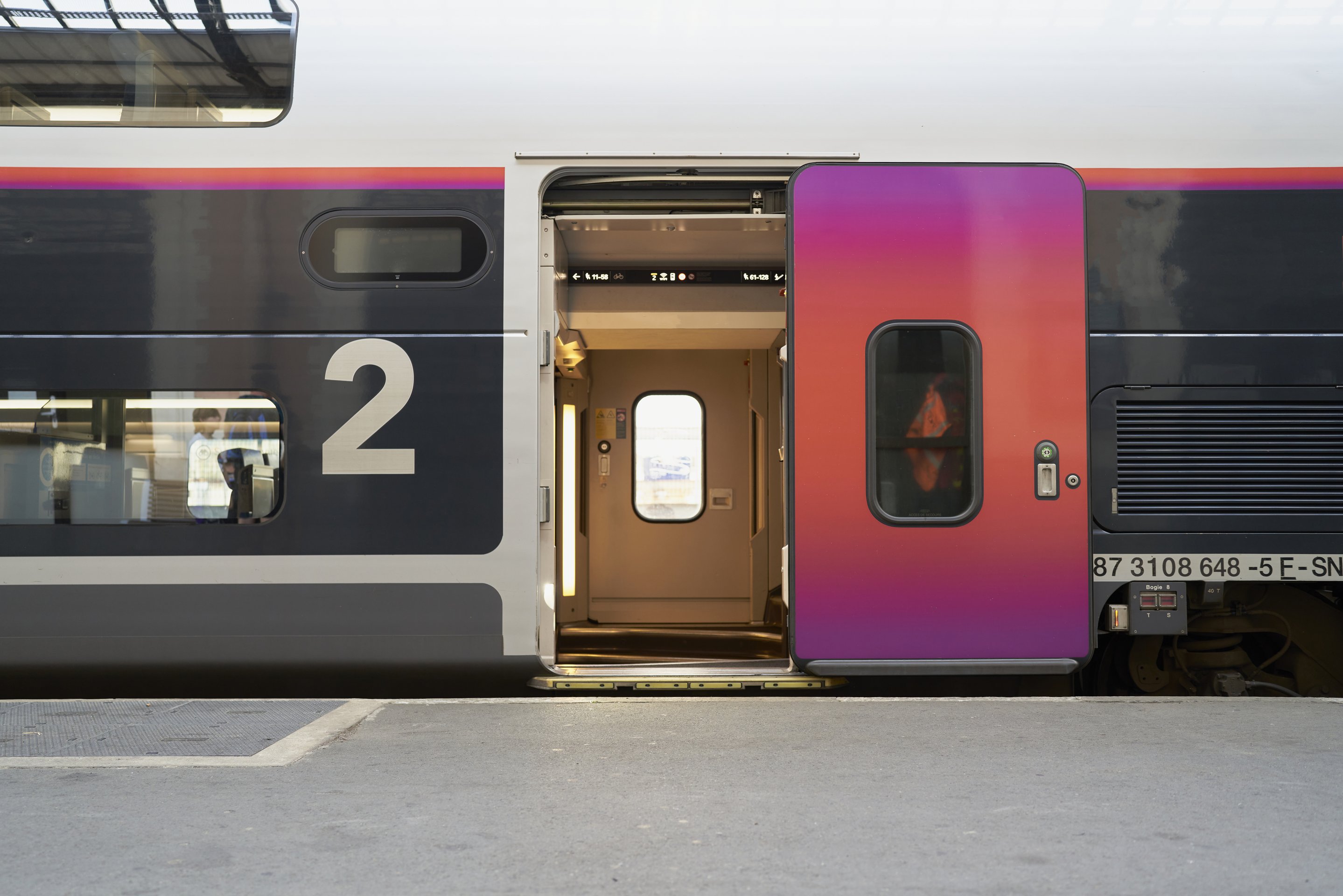 A side view of a double-decker passenger train carriage with an open, vibrant purple and orange door at a station platform.