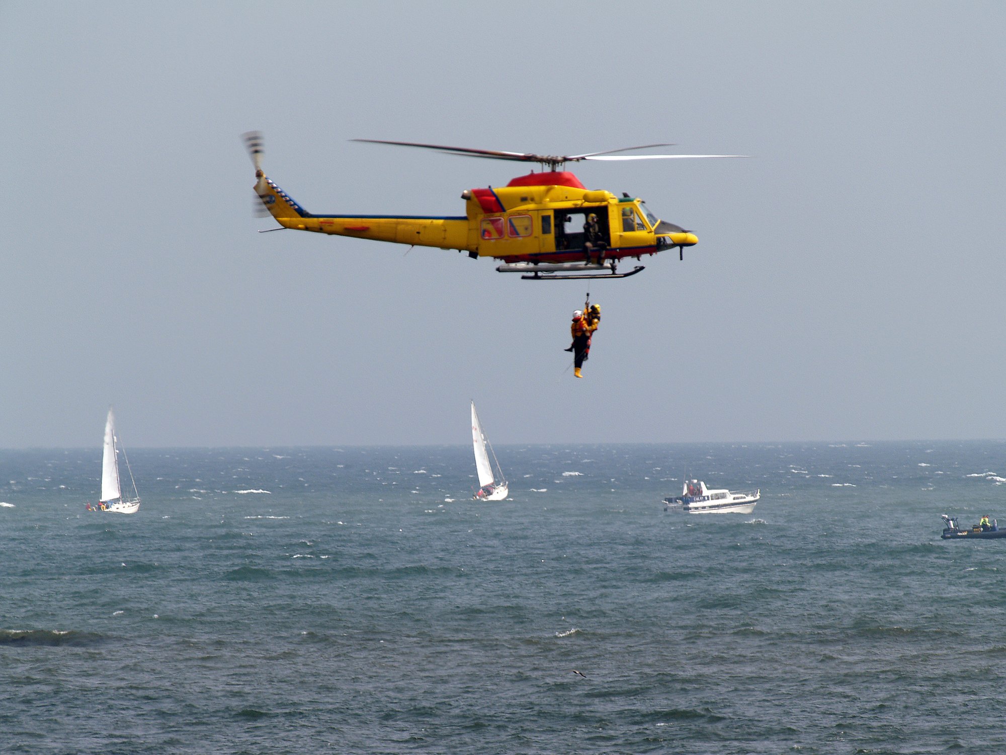 A search and rescue helicopter performs a hoist operation over the sea with sailboats in the background.