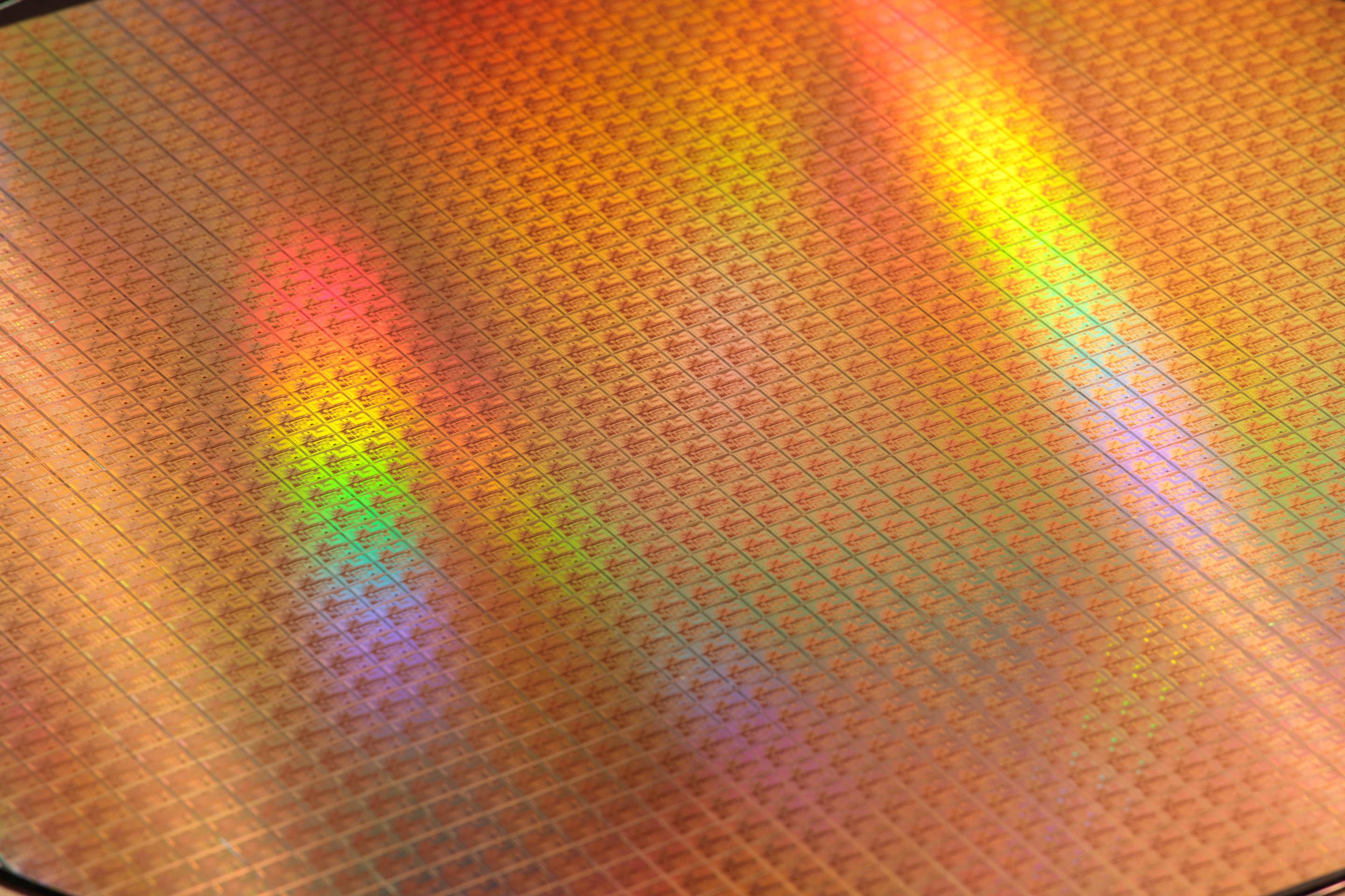 A high-angle close-up of a silicon wafer displaying an iridescent grid of microchip dies under reflective industrial lighting.