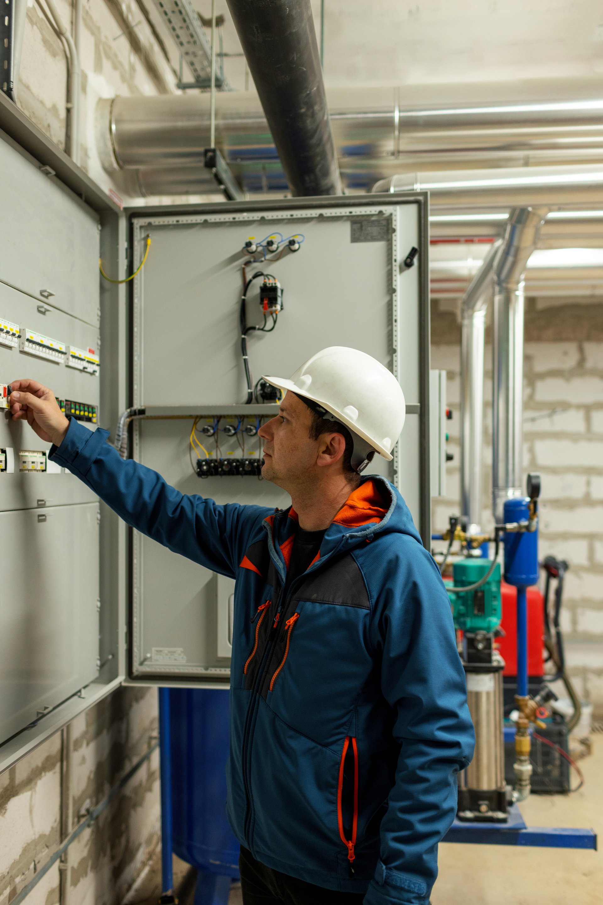 A professional technician in a hard hat inspects and operates an industrial electrical control panel inside a mechanical utility room.