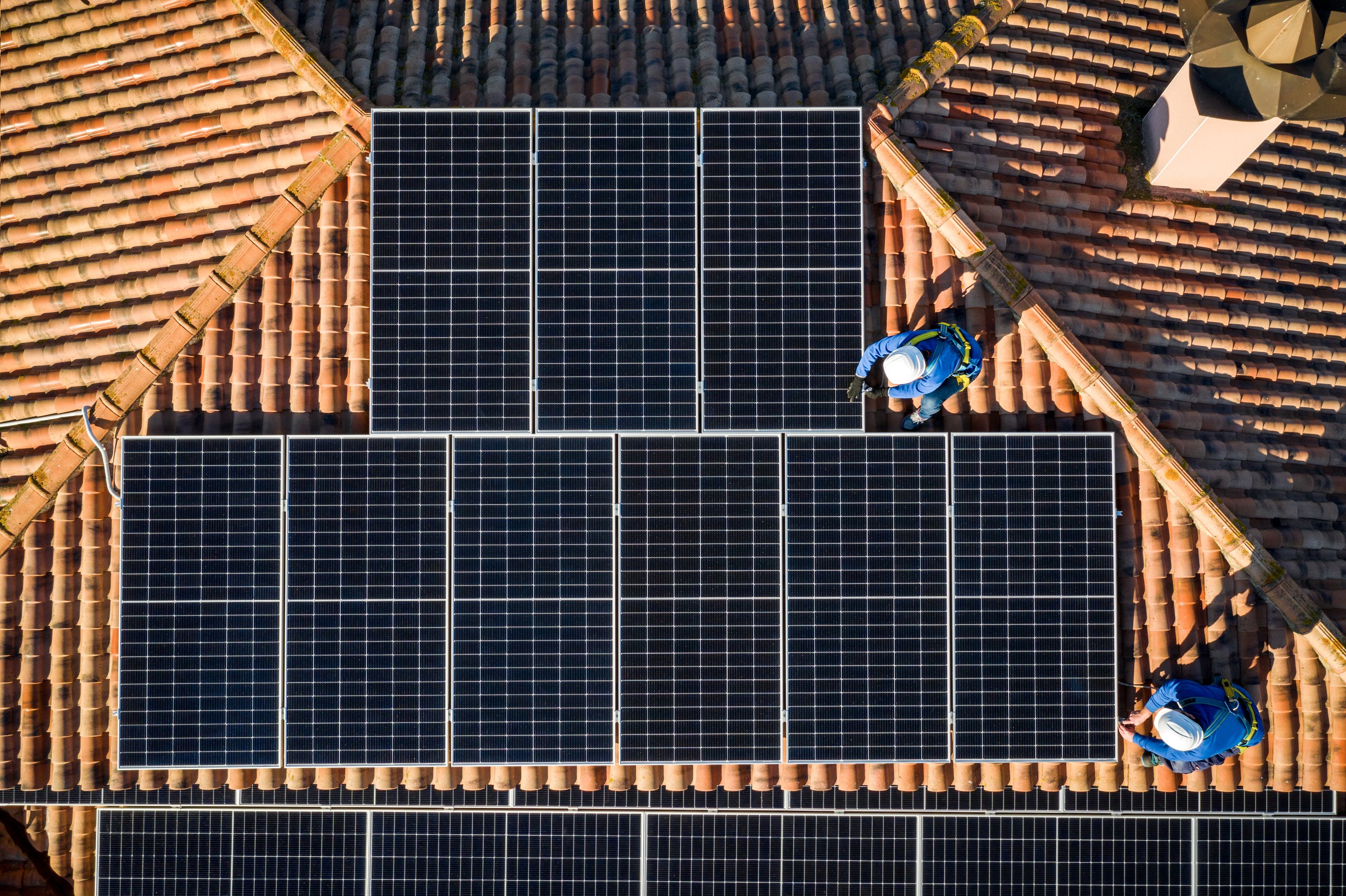 Aerial view of solar technicians installing photovoltaic panels on a traditional terracotta tile roof during a sunny day.