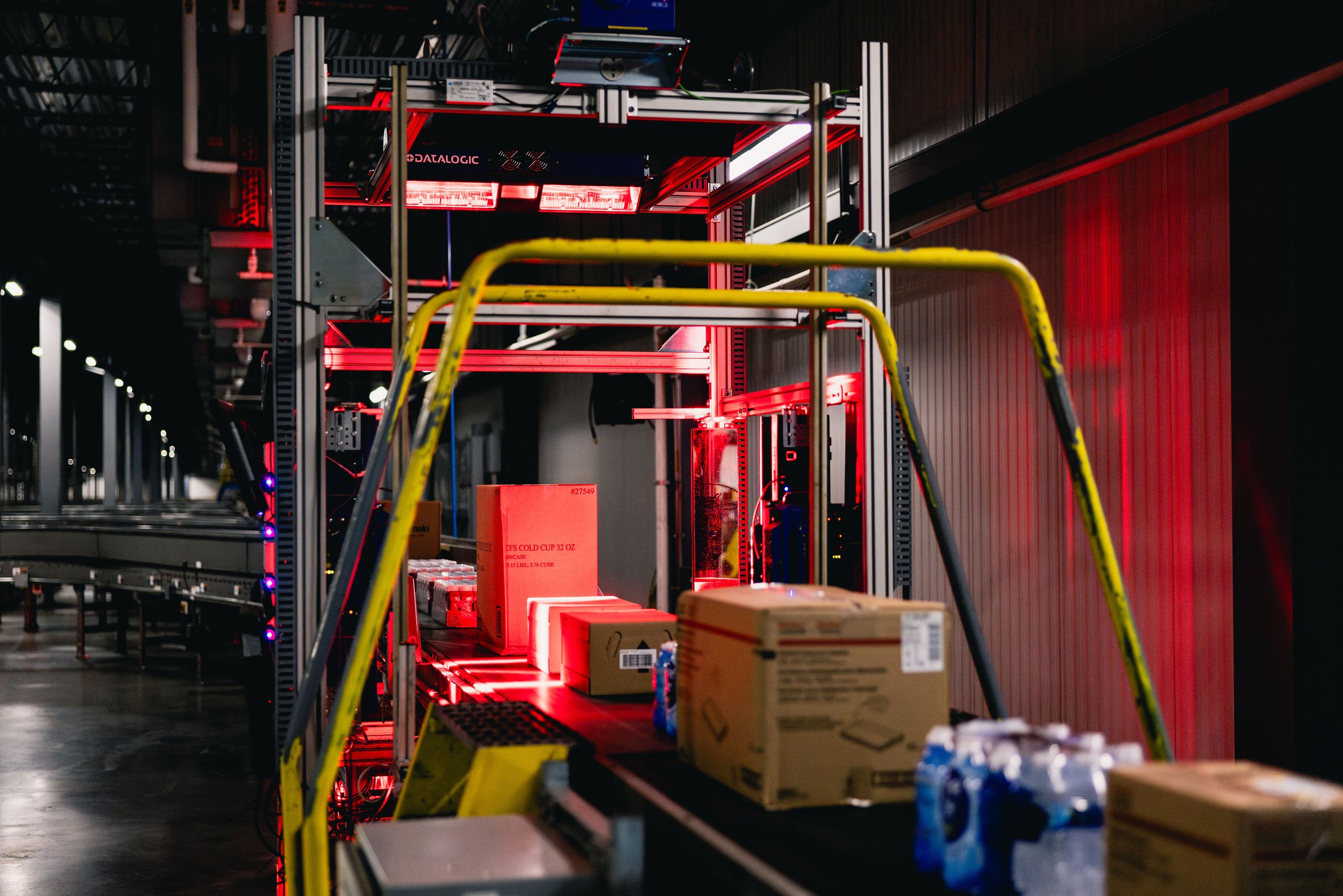 Cardboard boxes move along a conveyor belt under red industrial scanners for automated tracking in a fulfillment center.