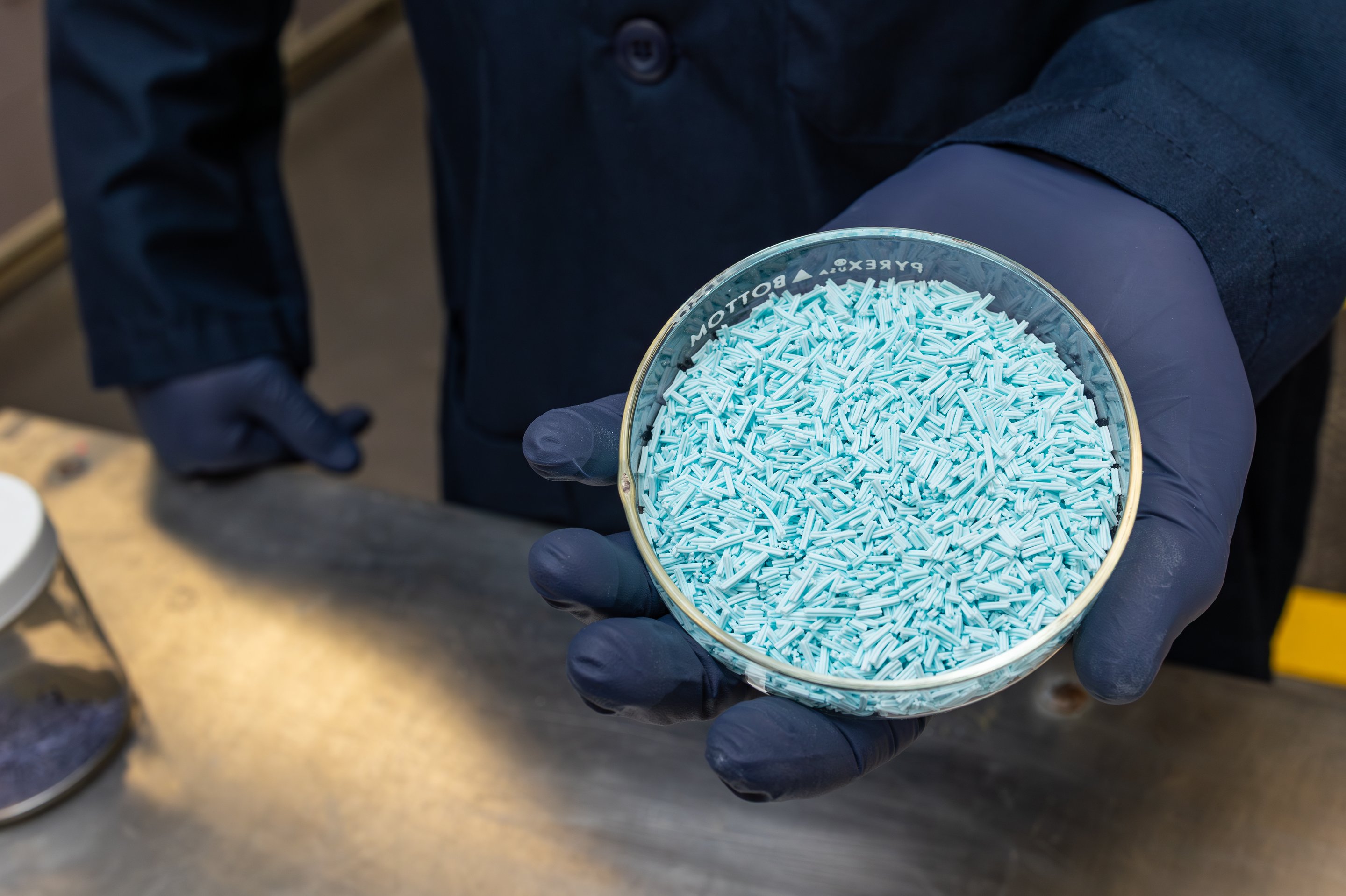 A technician in a lab coat and gloves holds a petri dish filled with light blue cylindrical chemical catalyst pellets for industrial quality inspection.