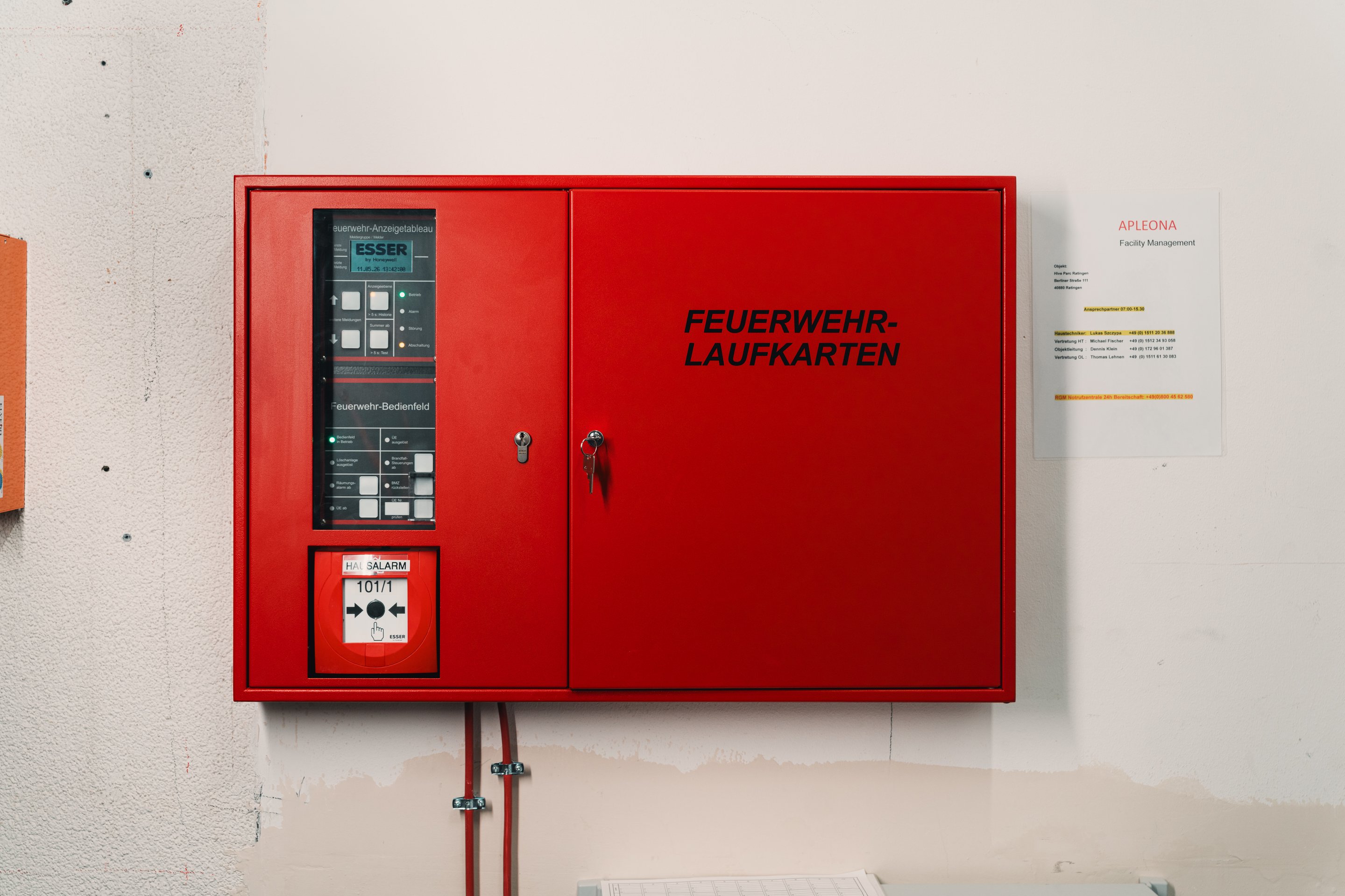 A red wall-mounted Esser fire alarm control panel with emergency map storage and contact information for facility management.