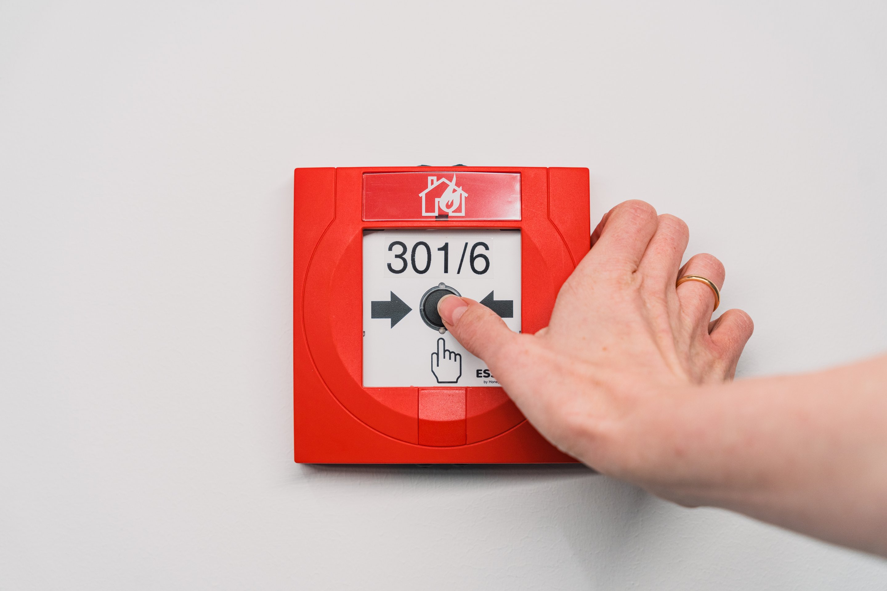 A person presses a red fire alarm call point button mounted on a white wall.