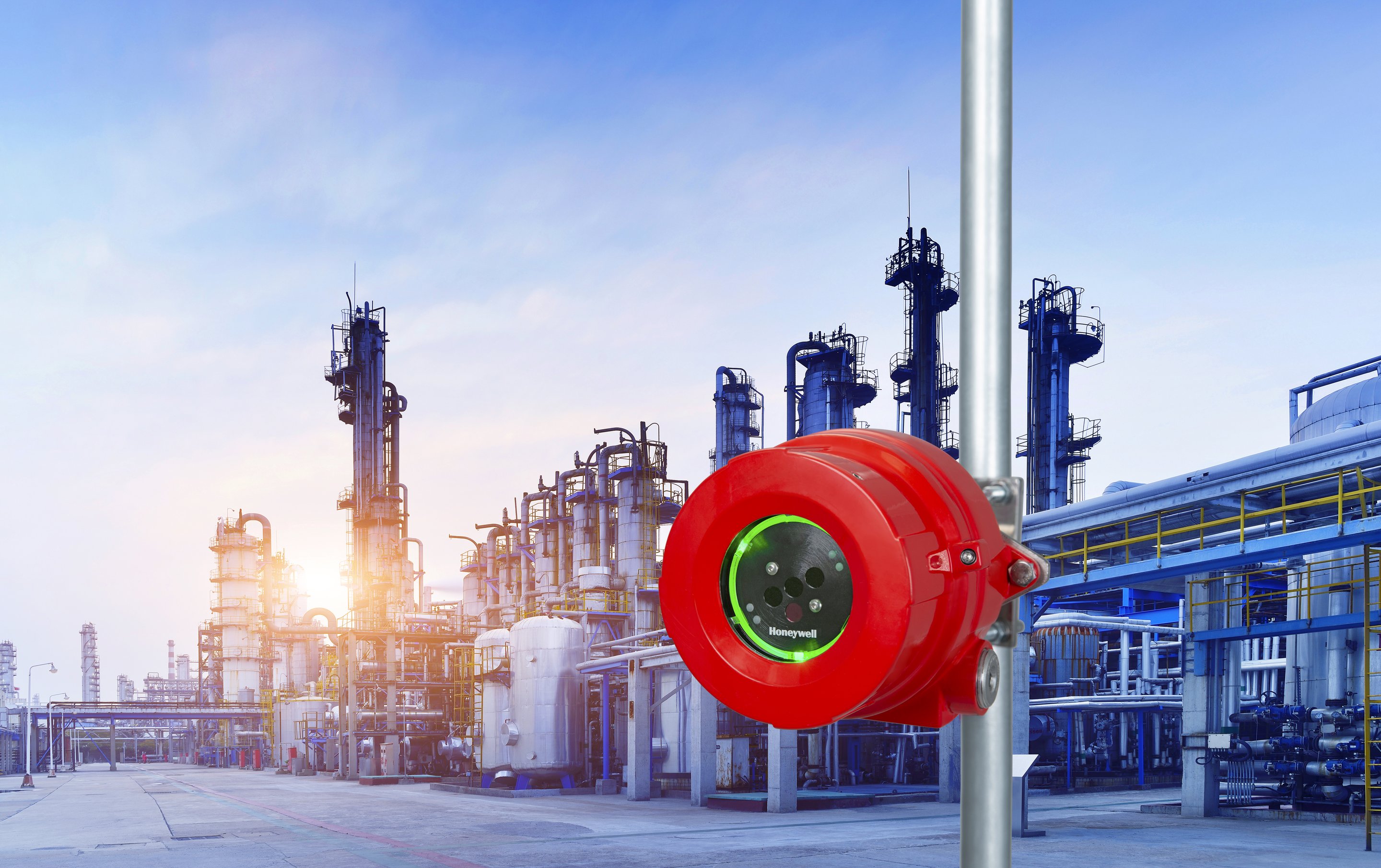 A bright red Honeywell gas detection sensor mounted on a pole against the backdrop of a petrochemical refinery facility at sunset.