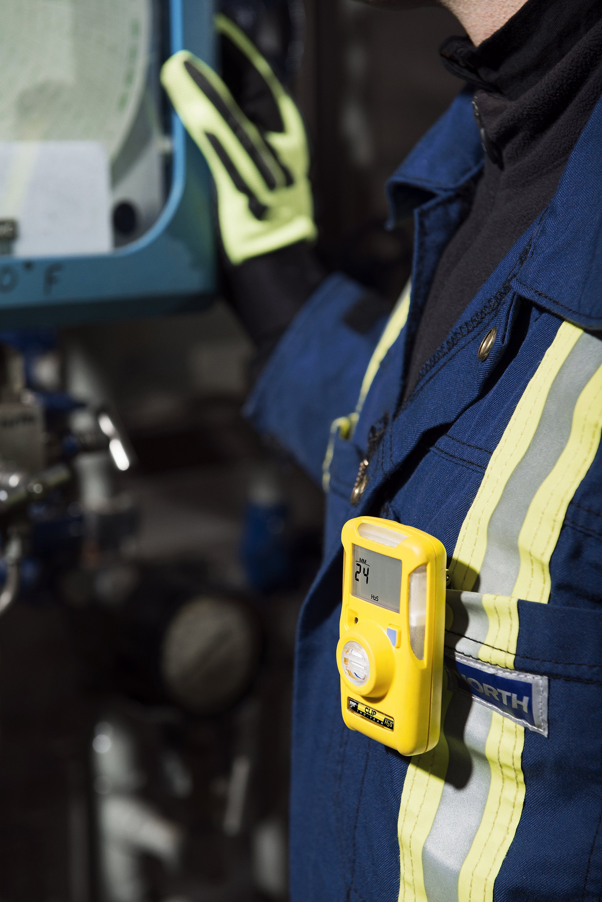 An industrial worker wears a personal hydrogen sulfide gas detector clipped to high-visibility work coveralls while operating equipment.