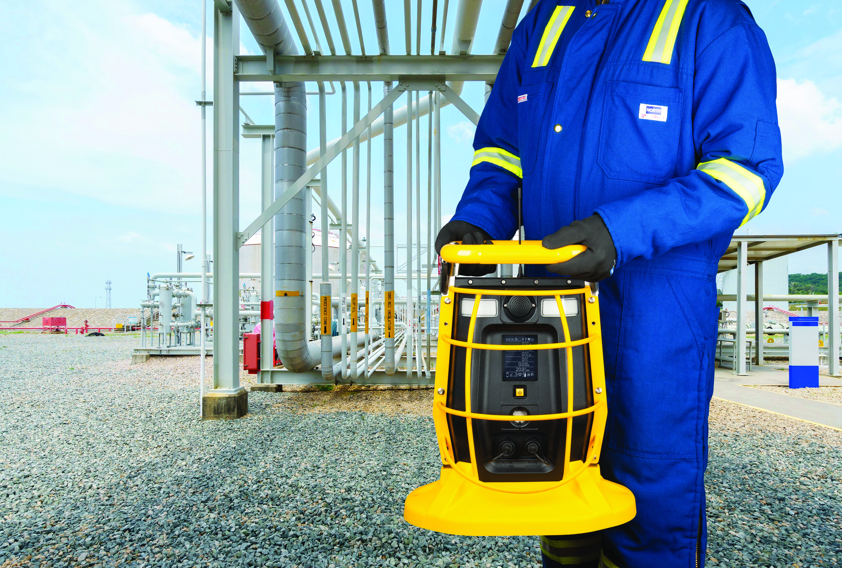 An industrial worker holds a portable gas detection monitor while standing at a processing facility featuring outdoor pipeline infrastructure.