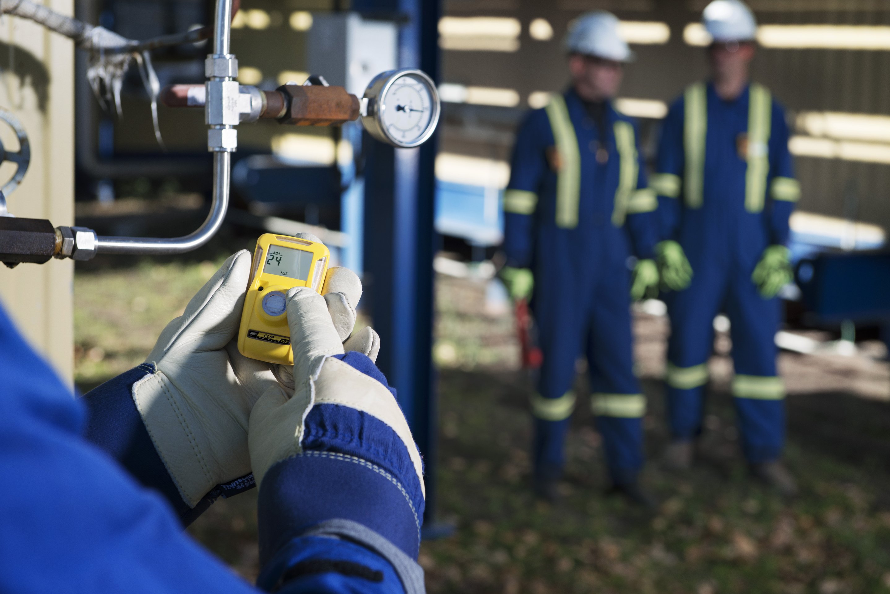 A worker in protective gear checks a portable gas detector near industrial piping, with colleagues standing blurred in the background.