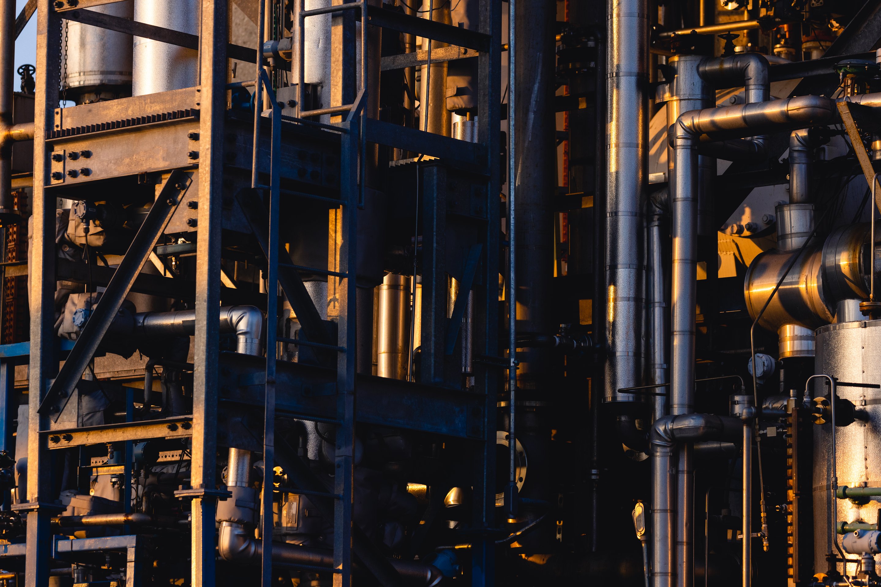 Sunlight illuminates metallic pipes and structural steel frames within a dense industrial refinery complex at dusk.