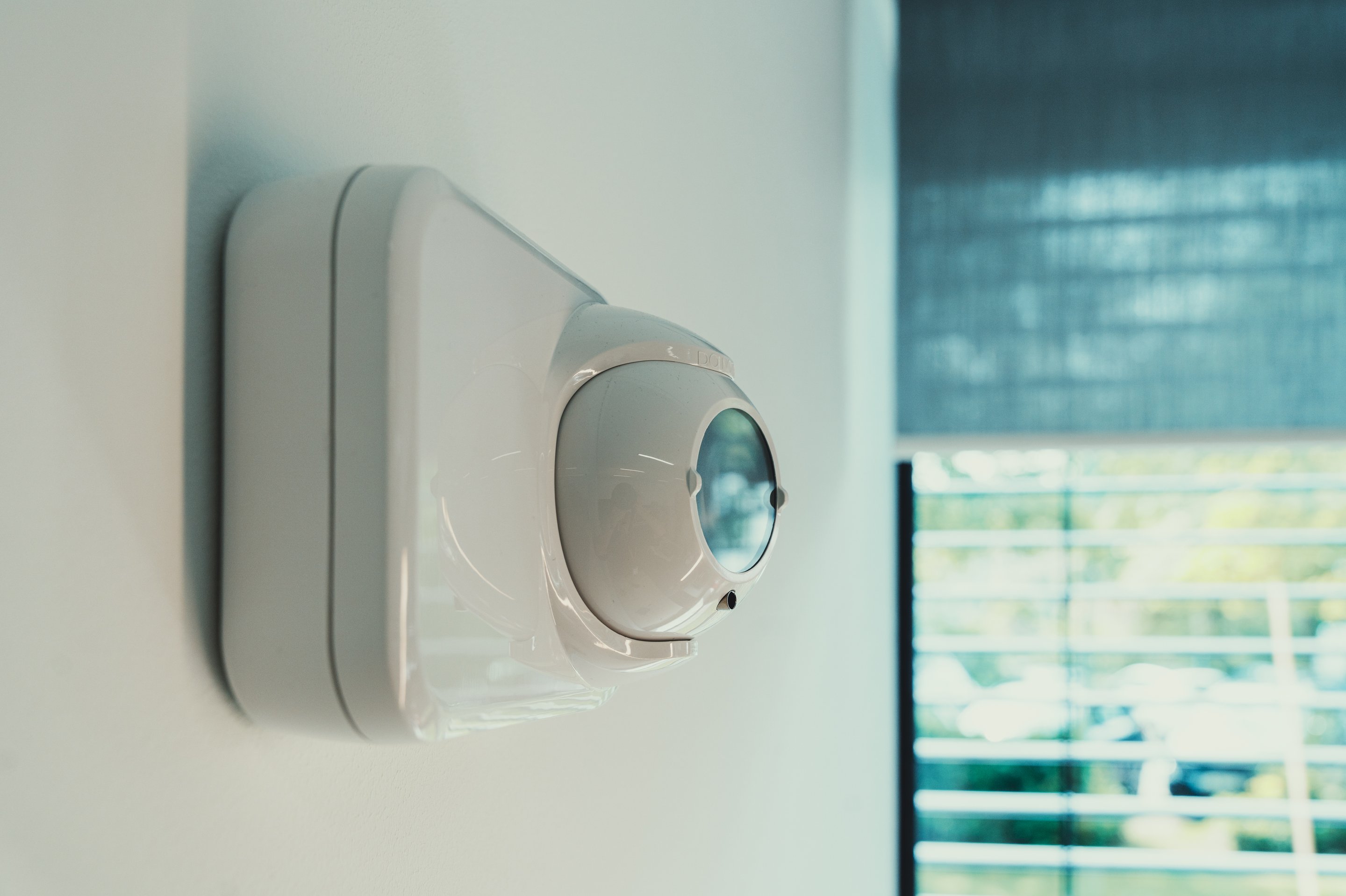A close-up side profile of a modern white motion detection sensor mounted on a clean office wall near a window.