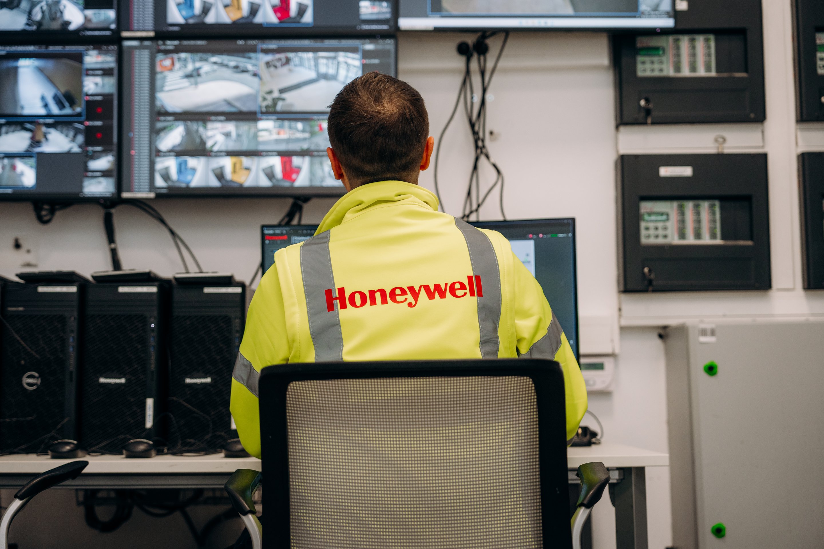 A technician in a high-visibility jacket monitors multiple surveillance screens within a corporate security control room.