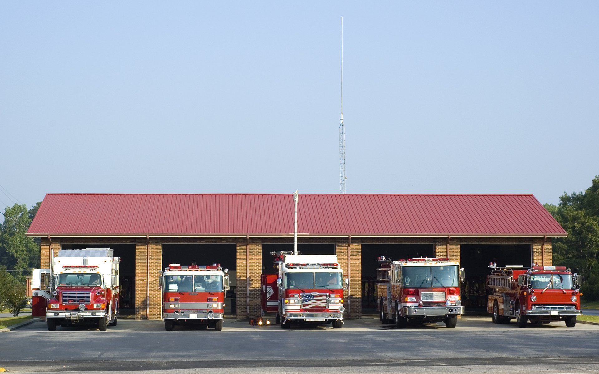 Four red fire engines parked in front of a brick fire station building under a clear blue sky.