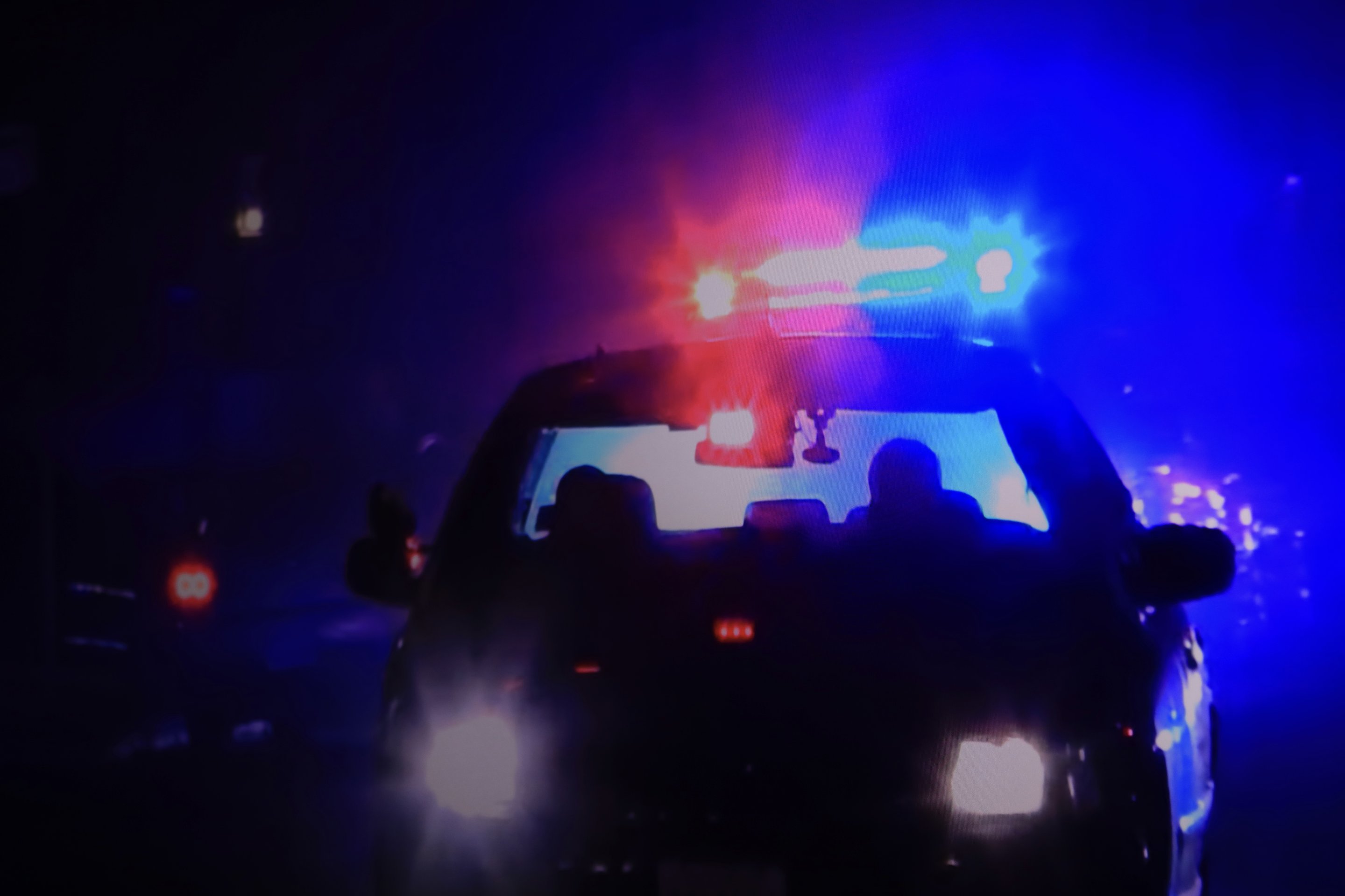 A police vehicle drives at night, illuminated by its bright, flashing red and blue emergency light bar.