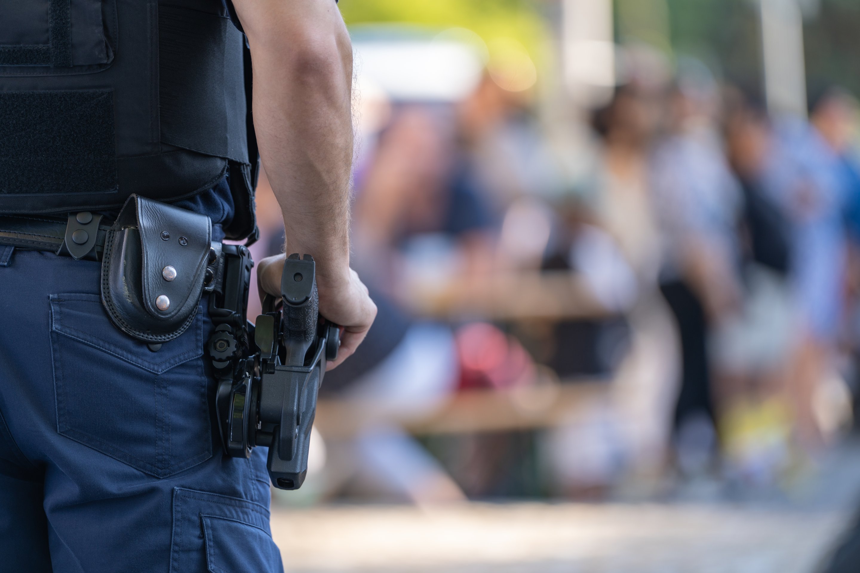 A police officer in tactical gear stands guard with their hand resting on a holstered sidearm against a blurred crowd background.