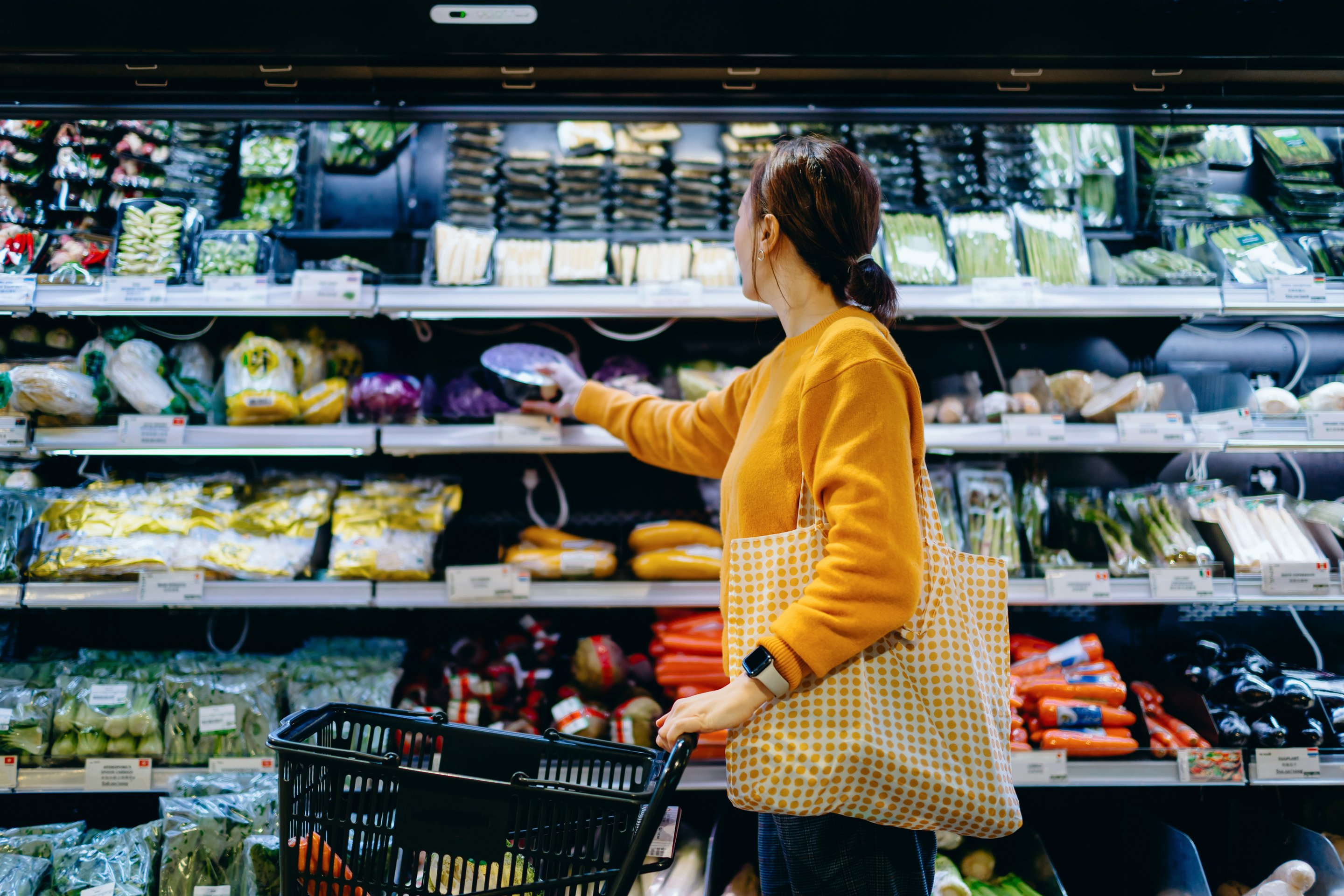A woman in a yellow sweater shops for fresh vegetables in a brightly lit, well-stocked grocery store aisle.