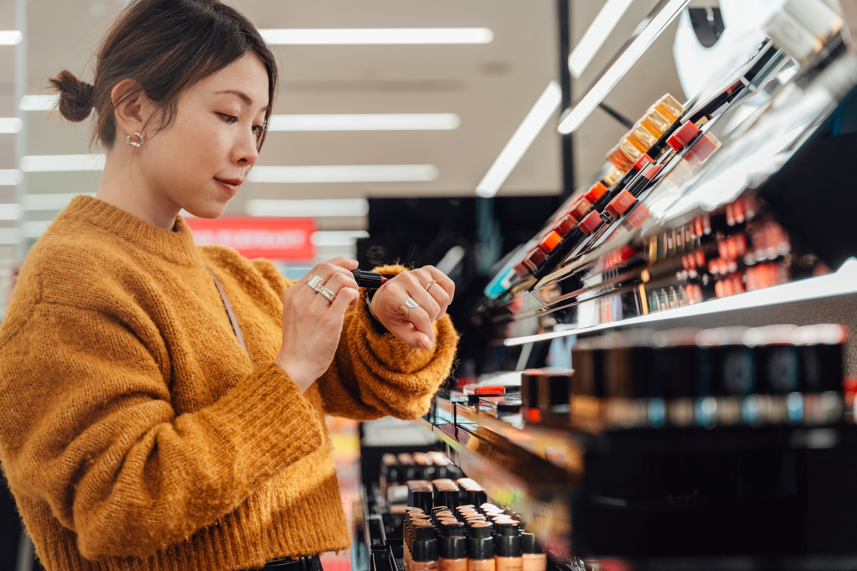 A focused woman in a knit sweater tests lipstick shade on her hand while shopping in a bright, modern cosmetics store.