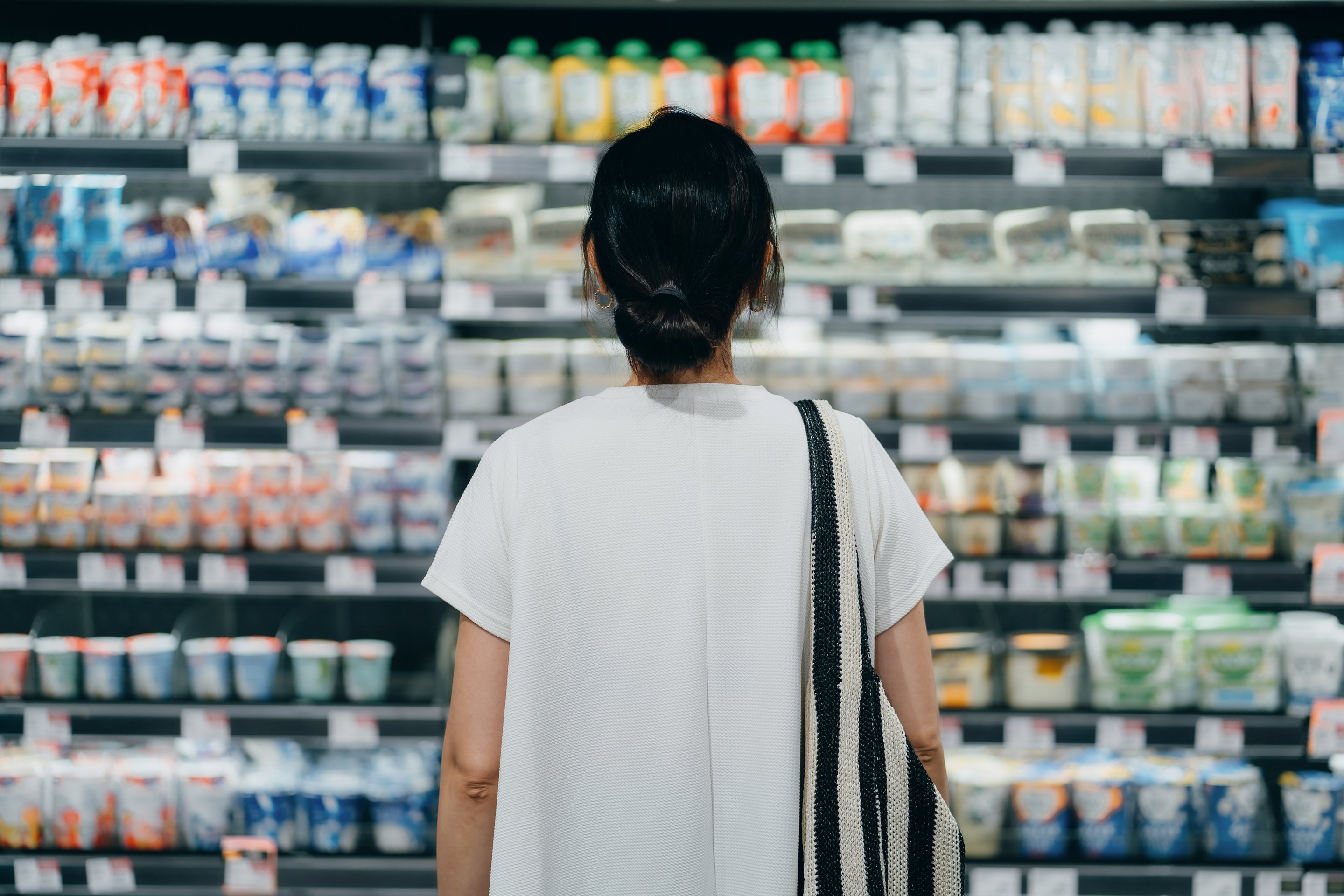 A woman with a shoulder bag stands before a well-stocked grocery store refrigerator shelf, carefully selecting dairy products.