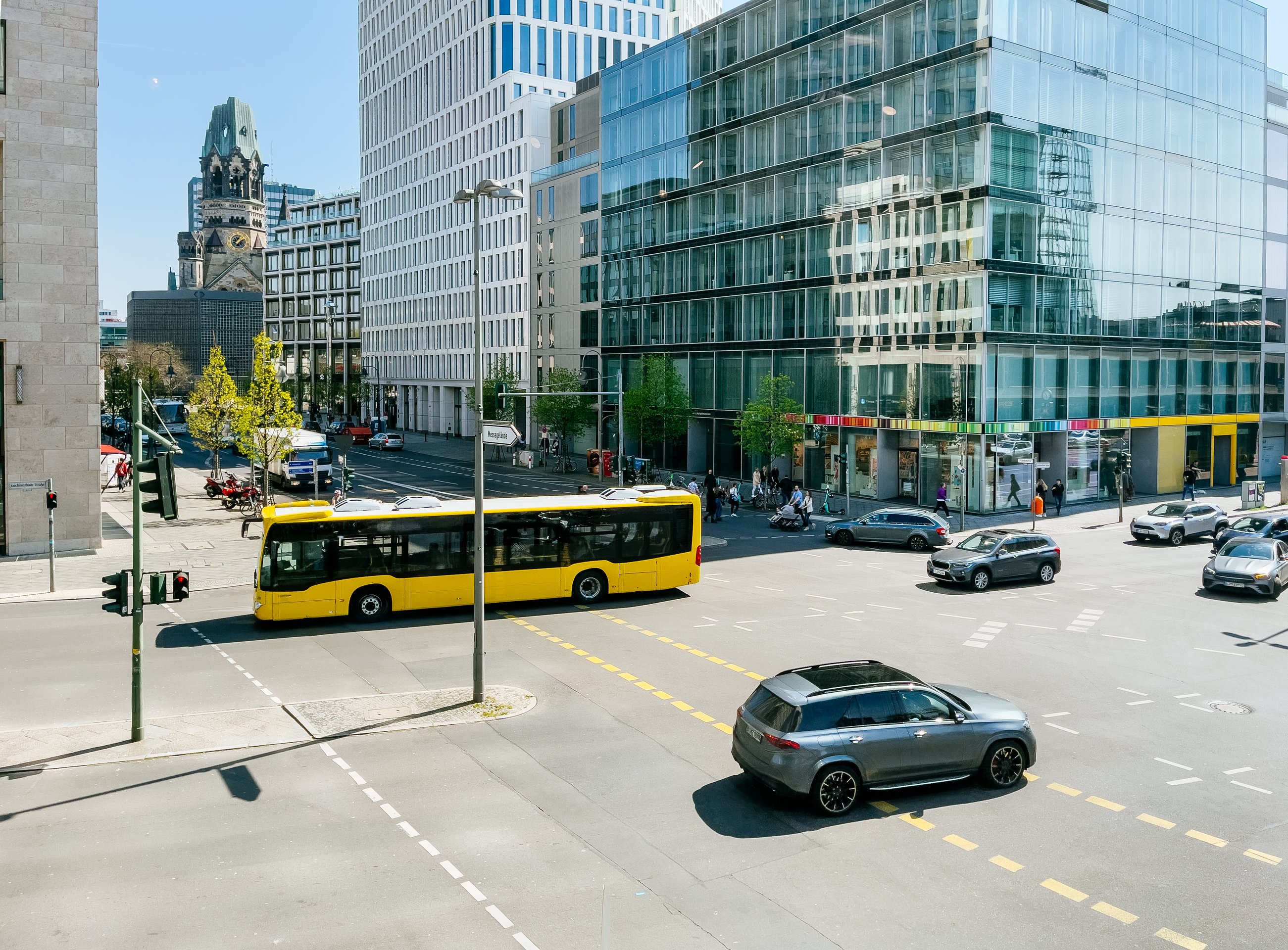 A bright yellow city bus and various cars navigate a busy Berlin street corner framed by modern glass architecture and the Kaiser Wilhelm Memorial Church.