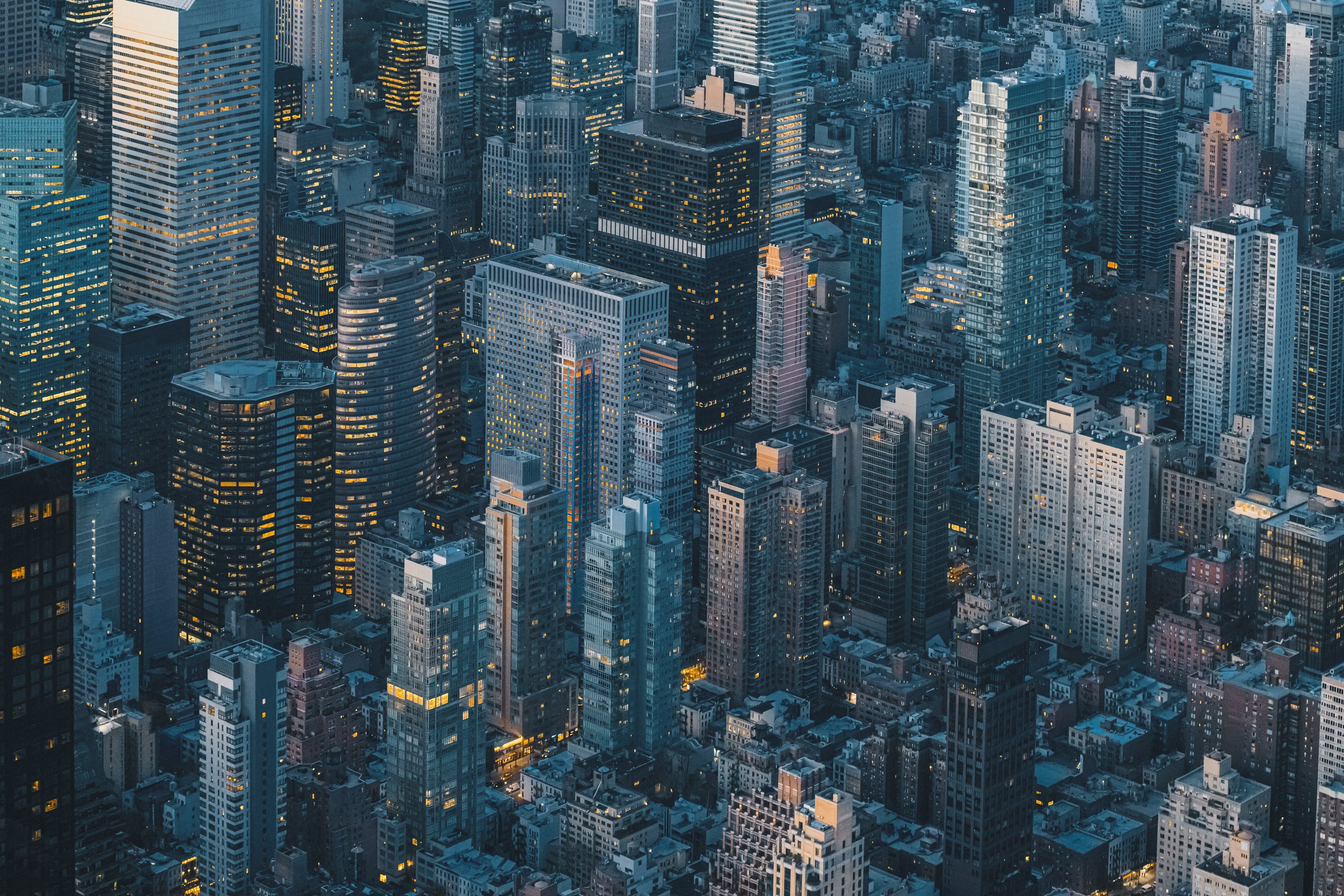 An aerial view of dense Manhattan skyscrapers illuminated by warm interior lighting against the cool blue tones of twilight.