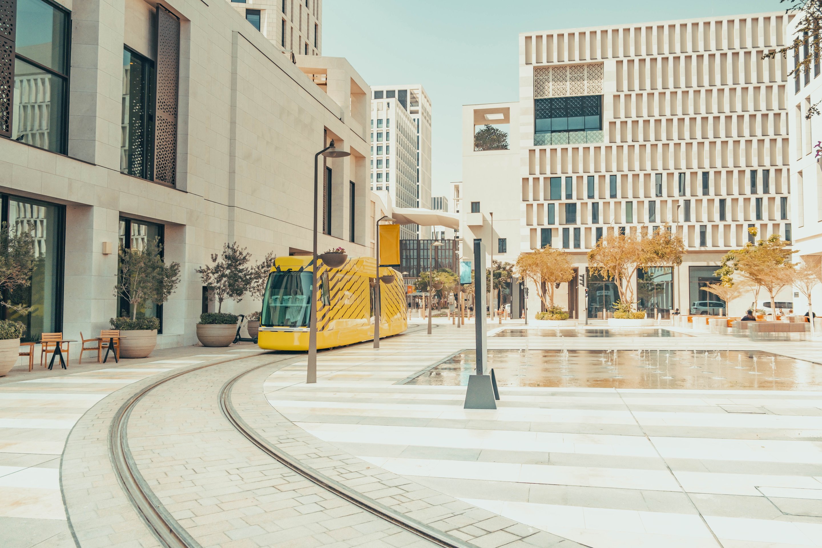 A modern yellow tram navigates tracks through a contemporary, sunlit urban plaza featuring geometric limestone buildings and a central water fountain.