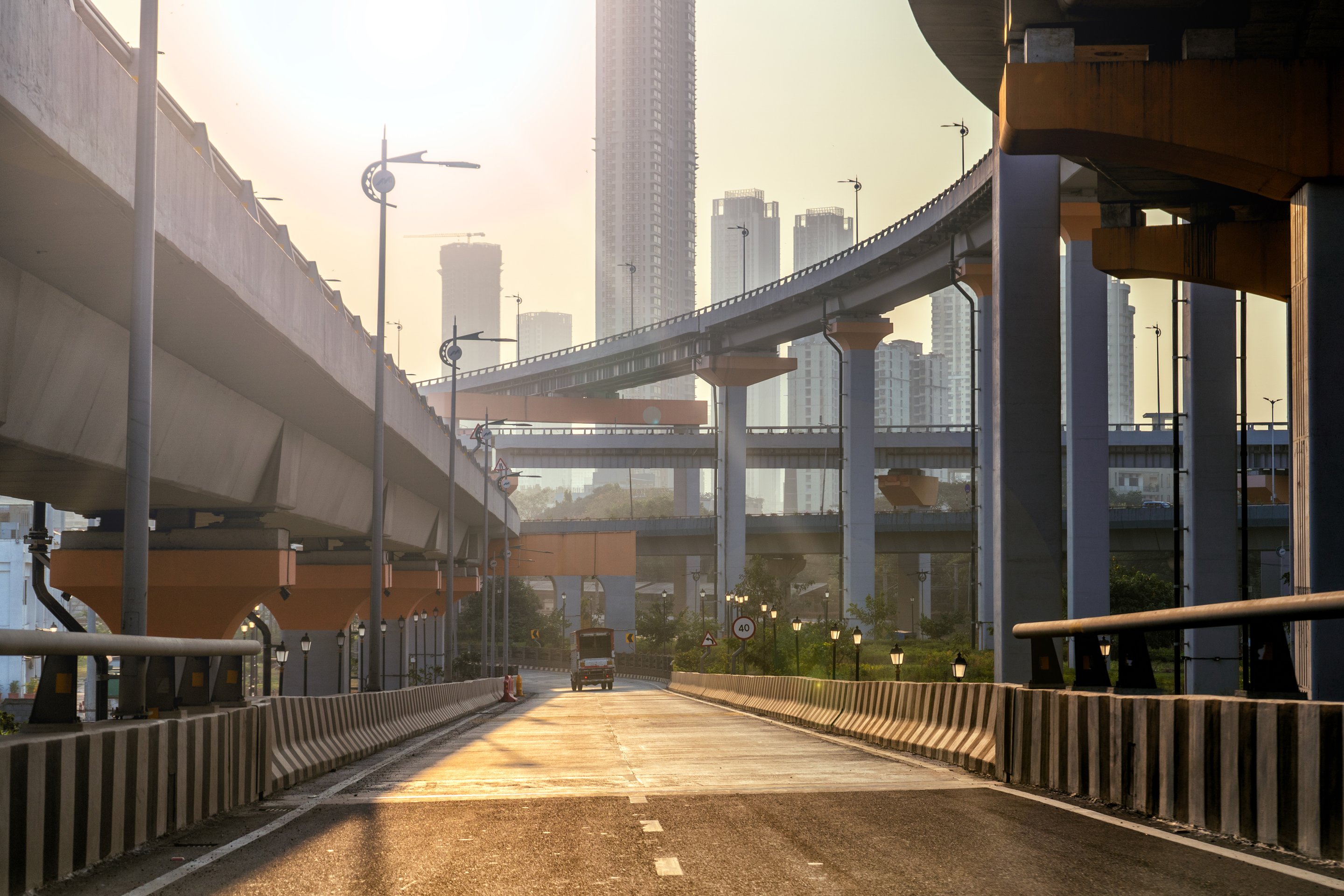 A single vehicle travels along a sunlit city highway beneath complex elevated multi-level interchanges against a backdrop of modern high-rise buildings.